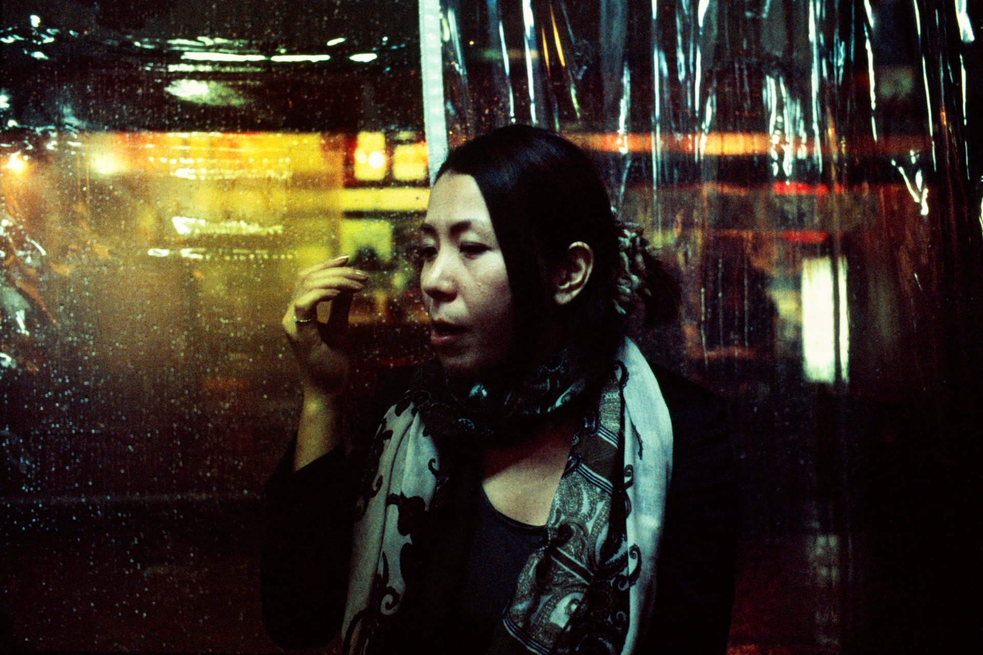 A woman with dark hair looks down as she presses her hand to a rain-covered window, with city and neon lights blurred in the background.