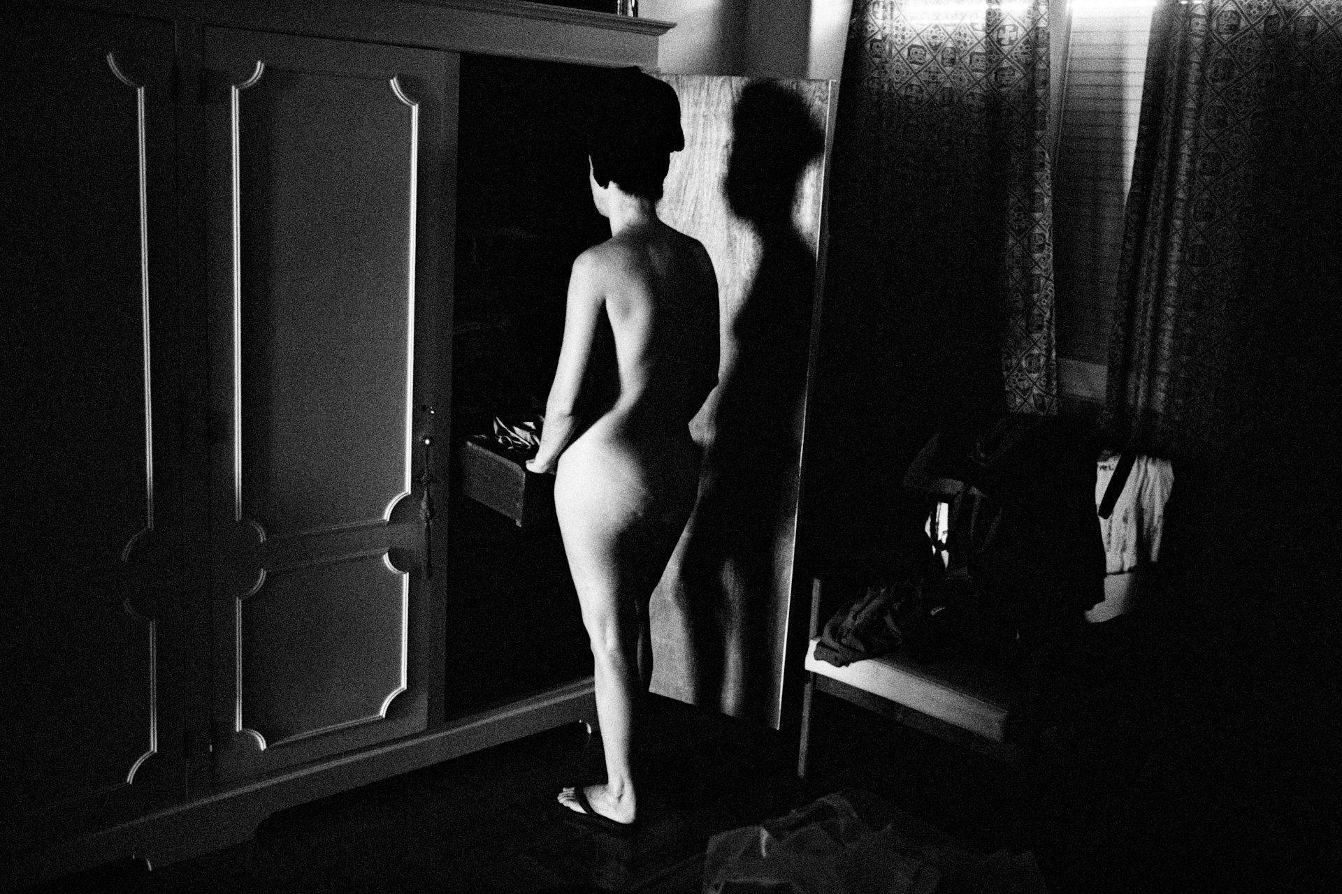 A black and white photo of a young person standing nude in a room, facing a mirror. Their back is turned to the camera, and their reflection can be seen in the mirror. The room has a wardrobe, a lamp, patterned curtains, and some clothes on a bench.