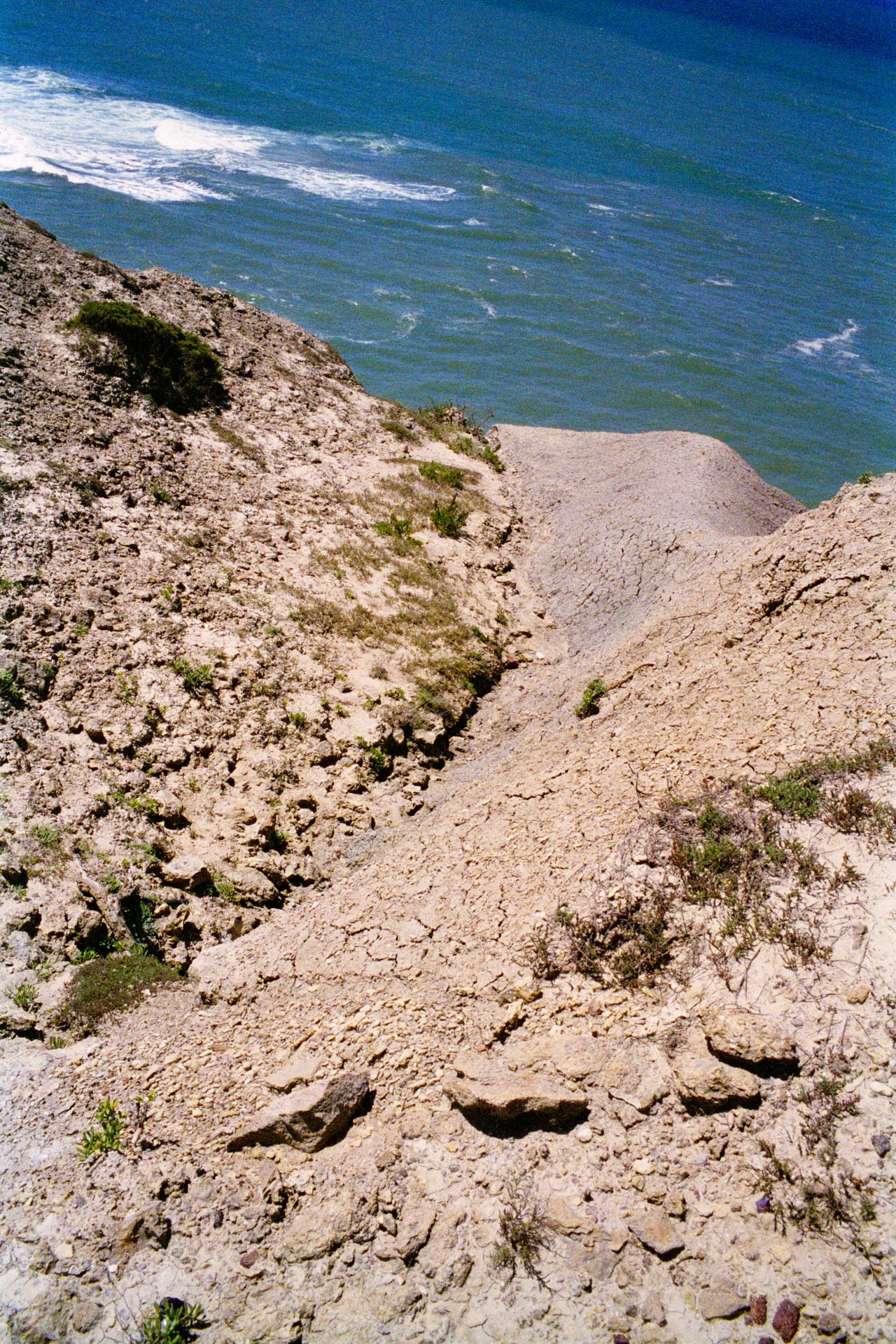 A steep, rocky hillside with sparse vegetation overlooking the ocean with blue water and white waves.
