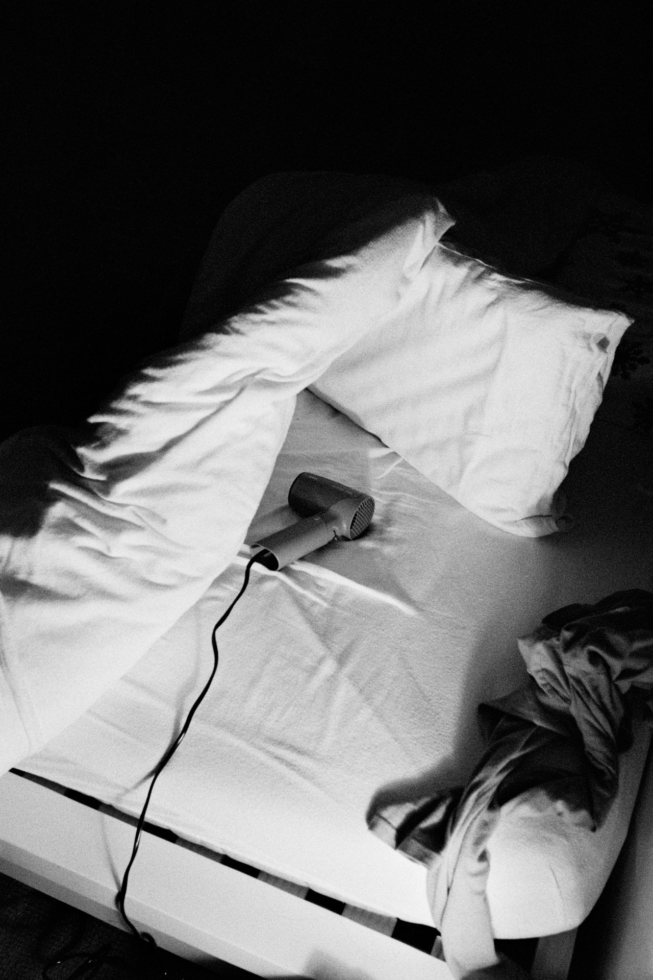 Unmade bed with white pillows, a crumpled blanket, and a hairdryer on the sheet in black and white.