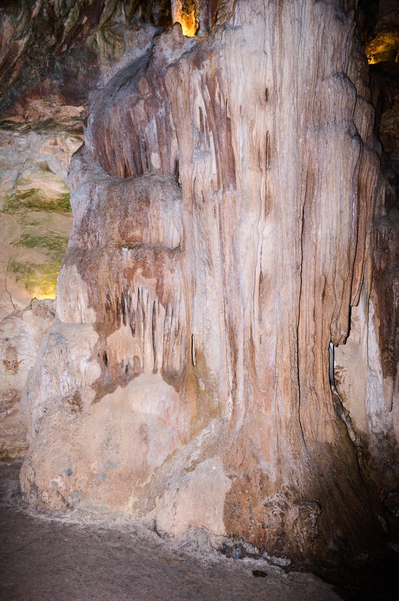 Interior view of a limestone cave with stalactites hanging from the ceiling and stalagmites rising from the ground, illuminated by warm artificial lighting.