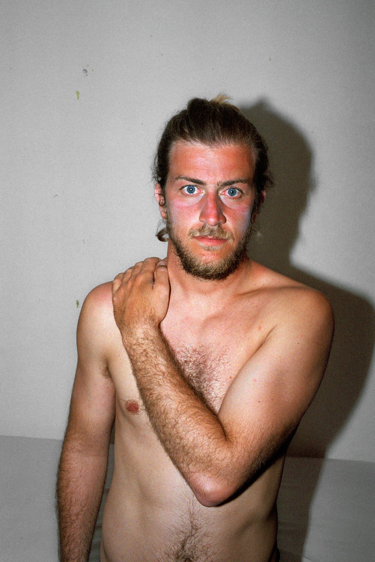 A topless man with long hair tied in a bun, blue eyes, a beard, and fair skin, standing against a plain wall, with his right hand touching his neck and shoulder.