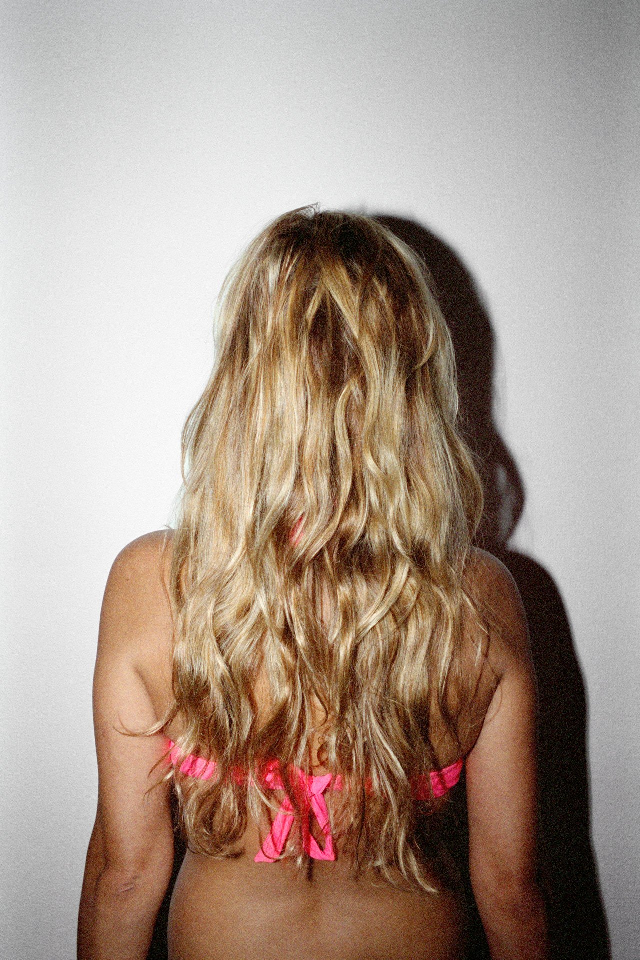 Back view of a woman with wavy blonde hair wearing a pink bikini top, standing against a plain white wall, casting a shadow.