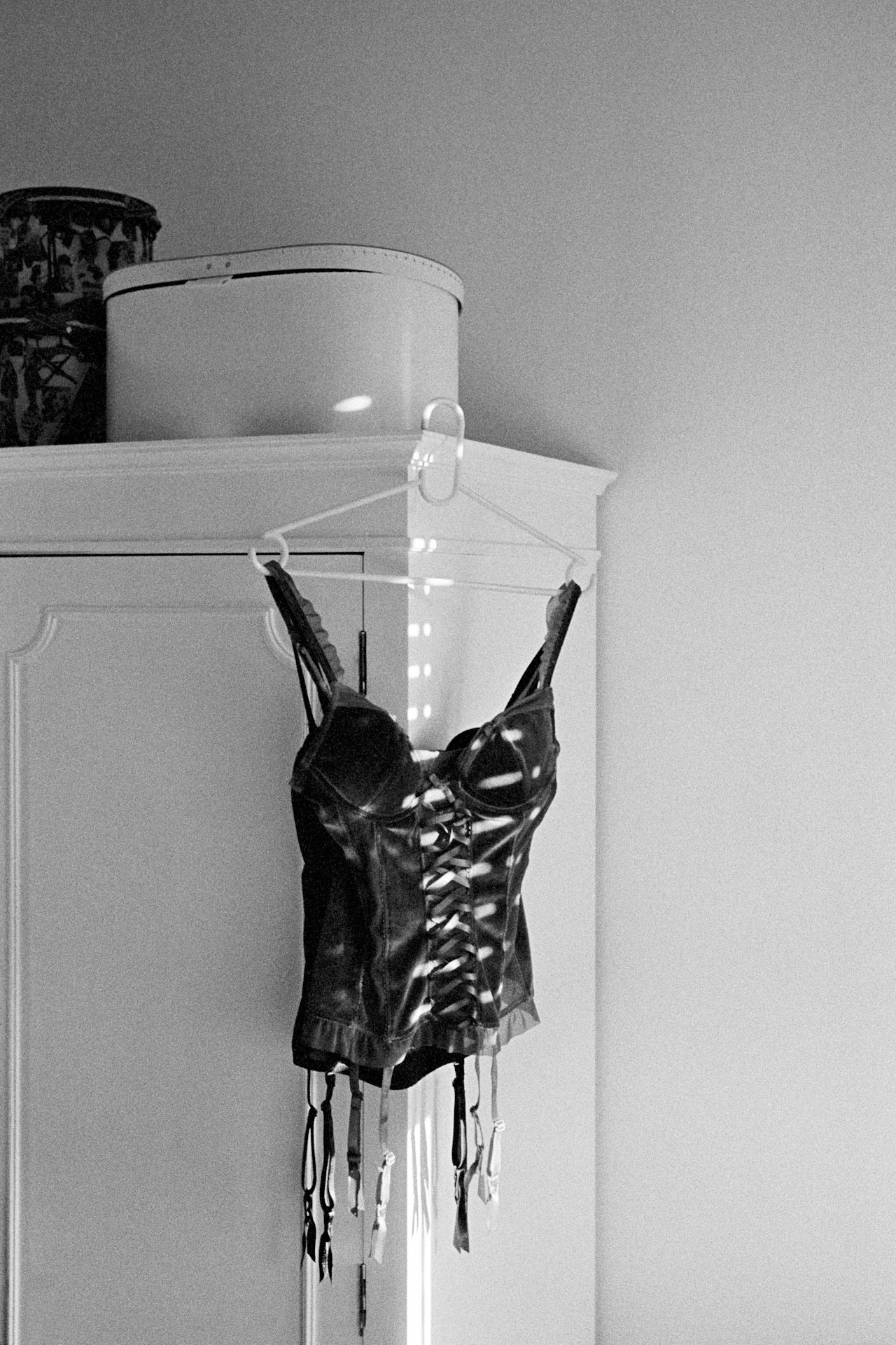 A black leather corset with lace-up front hanging from a clothesline inside a room, with a white cabinet and a round storage container on top of it.
