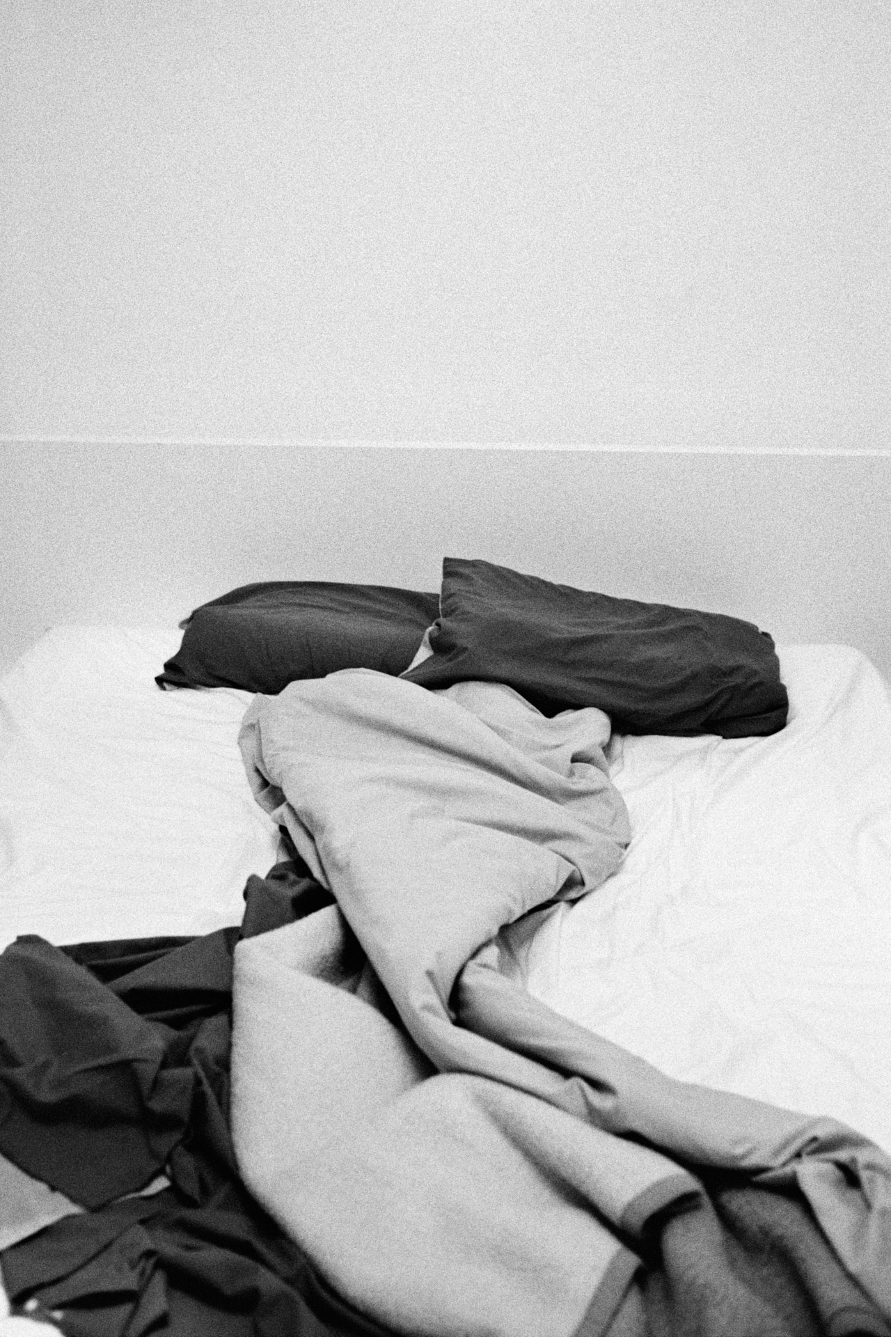 Messy bed with crumpled and disorganized sheets and blankets against a plain, light-colored wall.