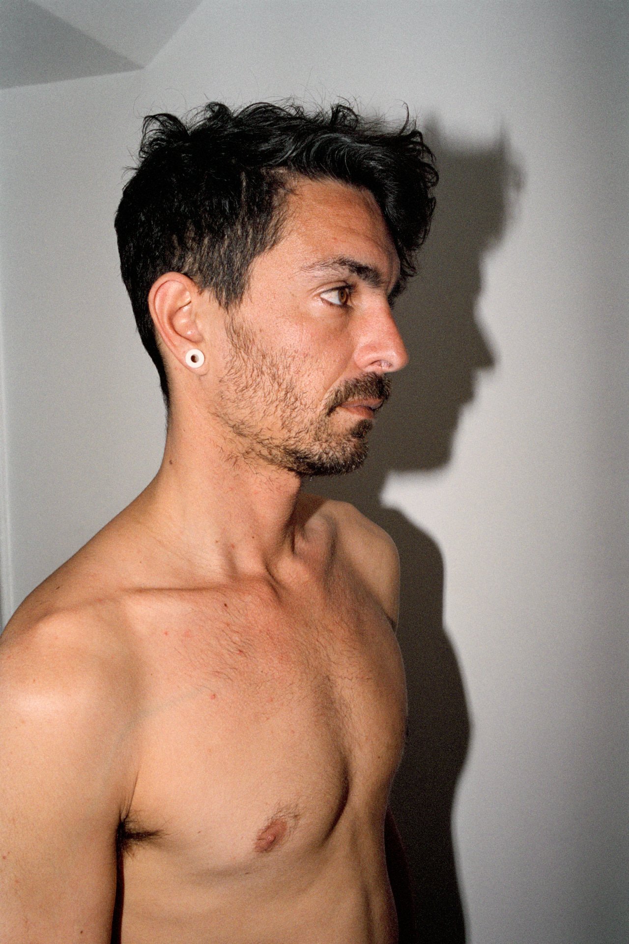 A shirtless young man with dark, curly hair, a beard, and single earrings, facing to the right with a shadow on the wall behind him.