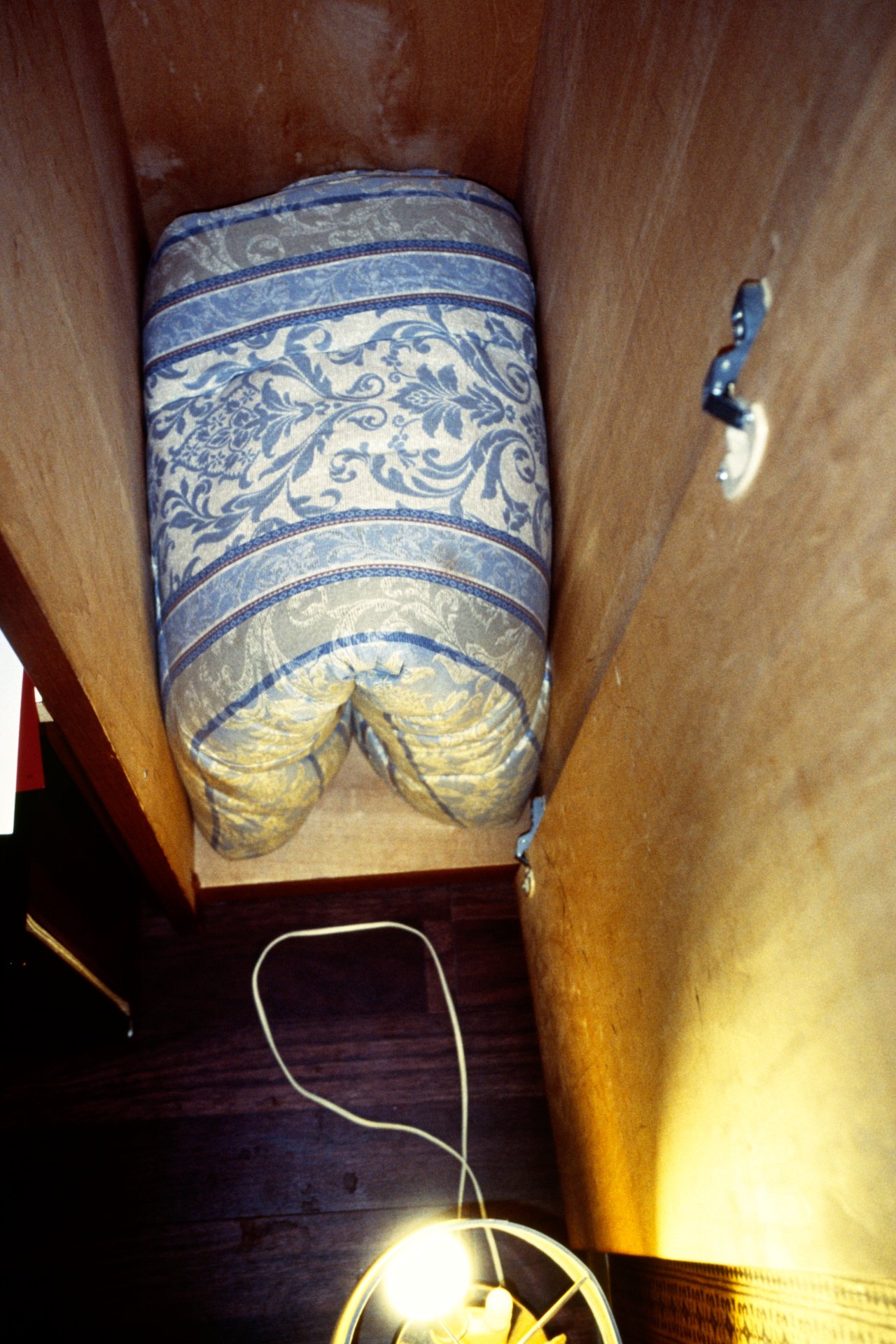 An open wooden cabinet showing a fuzzy blue and yellow patterned mattress stored inside. A yellow electrical cord is on the dark wooden floor beneath the cabinet, with a bright light fixture at the bottom.