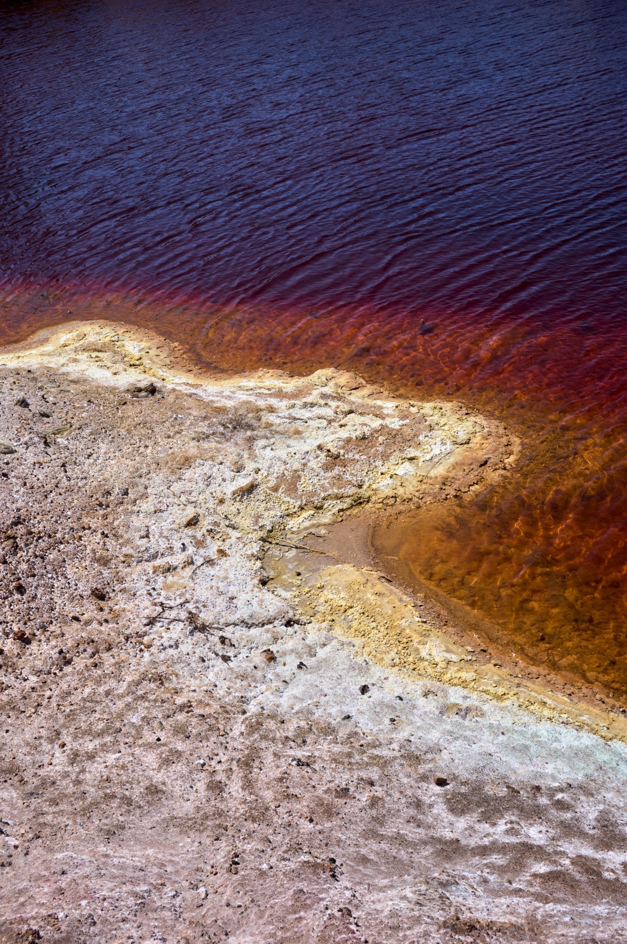 A shoreline with brown, cracked earth and a transition to a reddish water body, possibly a hot spring or mineral-rich lake.