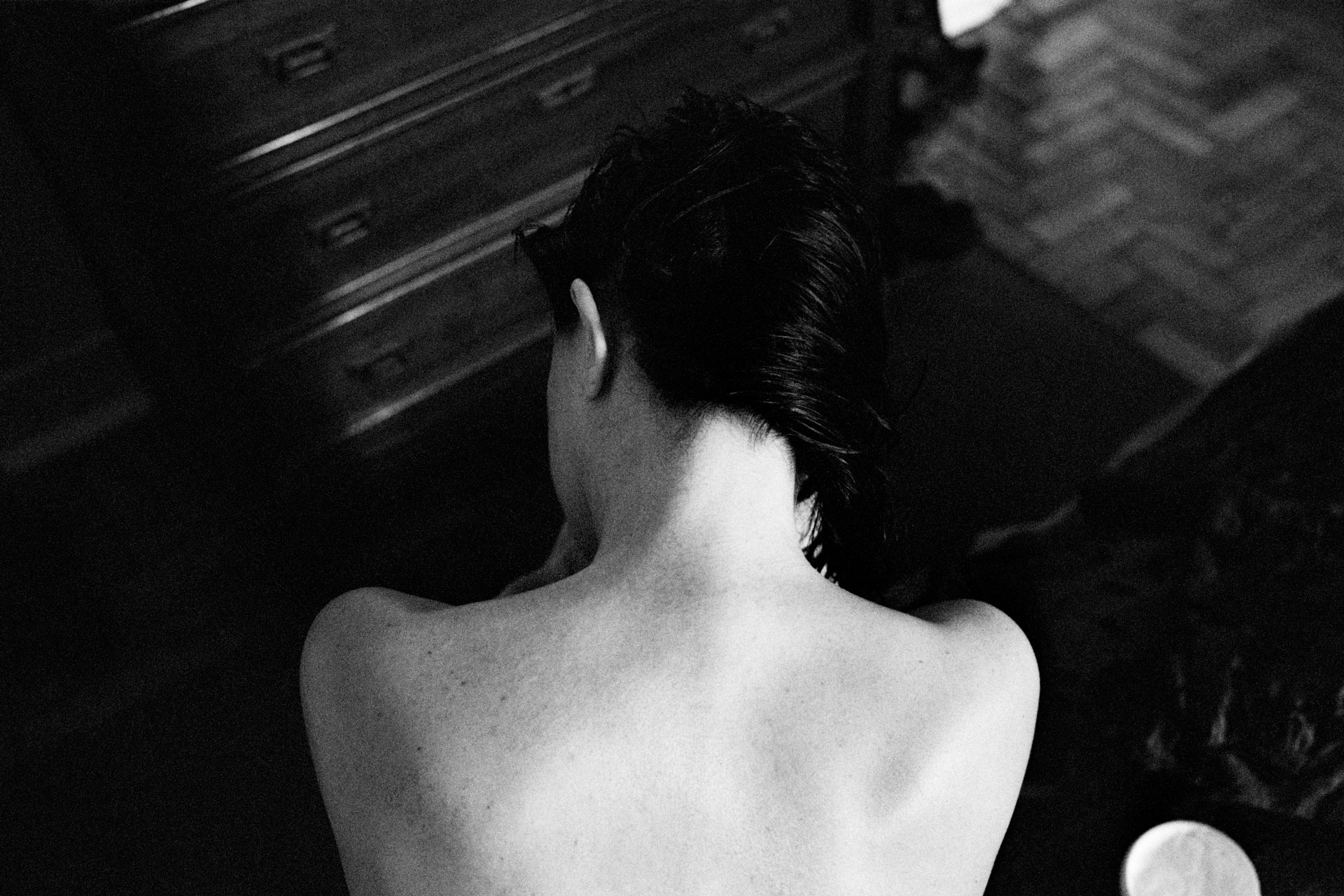 Back view of a shirtless person with wet, dark hair, facing a wooden dresser in a dimly lit room.