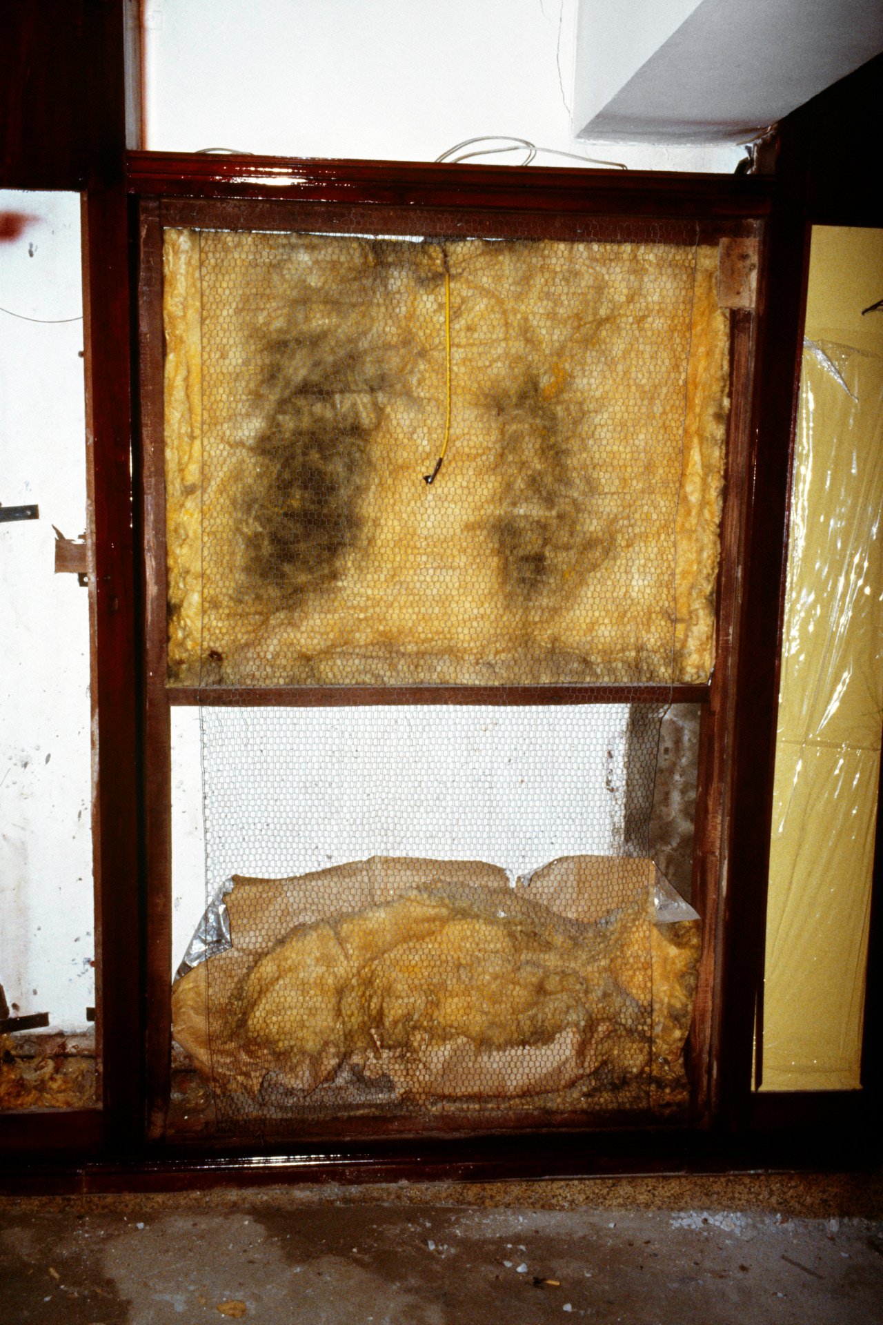 A wooden frame with honeycomb and insulation material, covered partly with wire mesh and plastic. The frame appears to be an unfinished or old honey extractor or hive structure.
