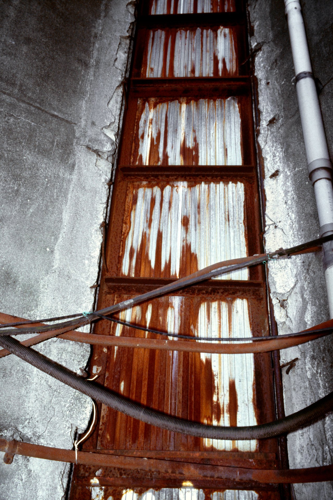 Rusty metal door with peeling paint, mounted on a concrete wall, with pipes and cables nearby.