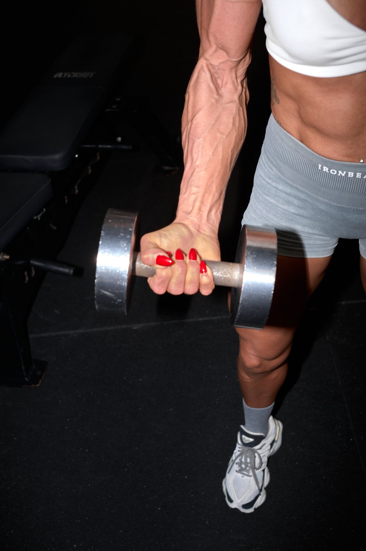 Person lifting a dumbbell in a gym, wearing a white tank top, grey shorts, and white sneakers.