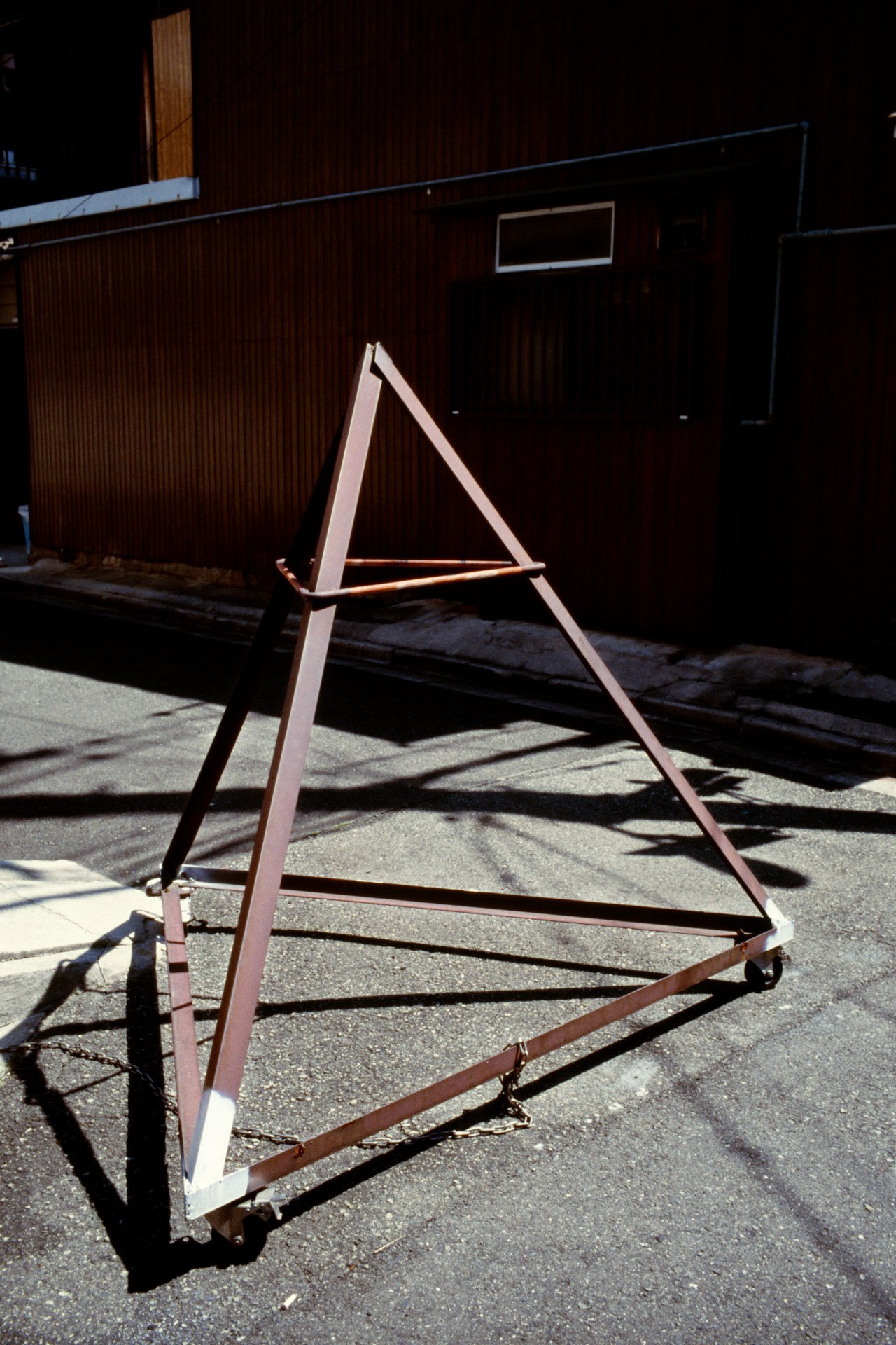 Empty metal A-frame sign stand on city street, casting shadow on pavement, with brown building in background.