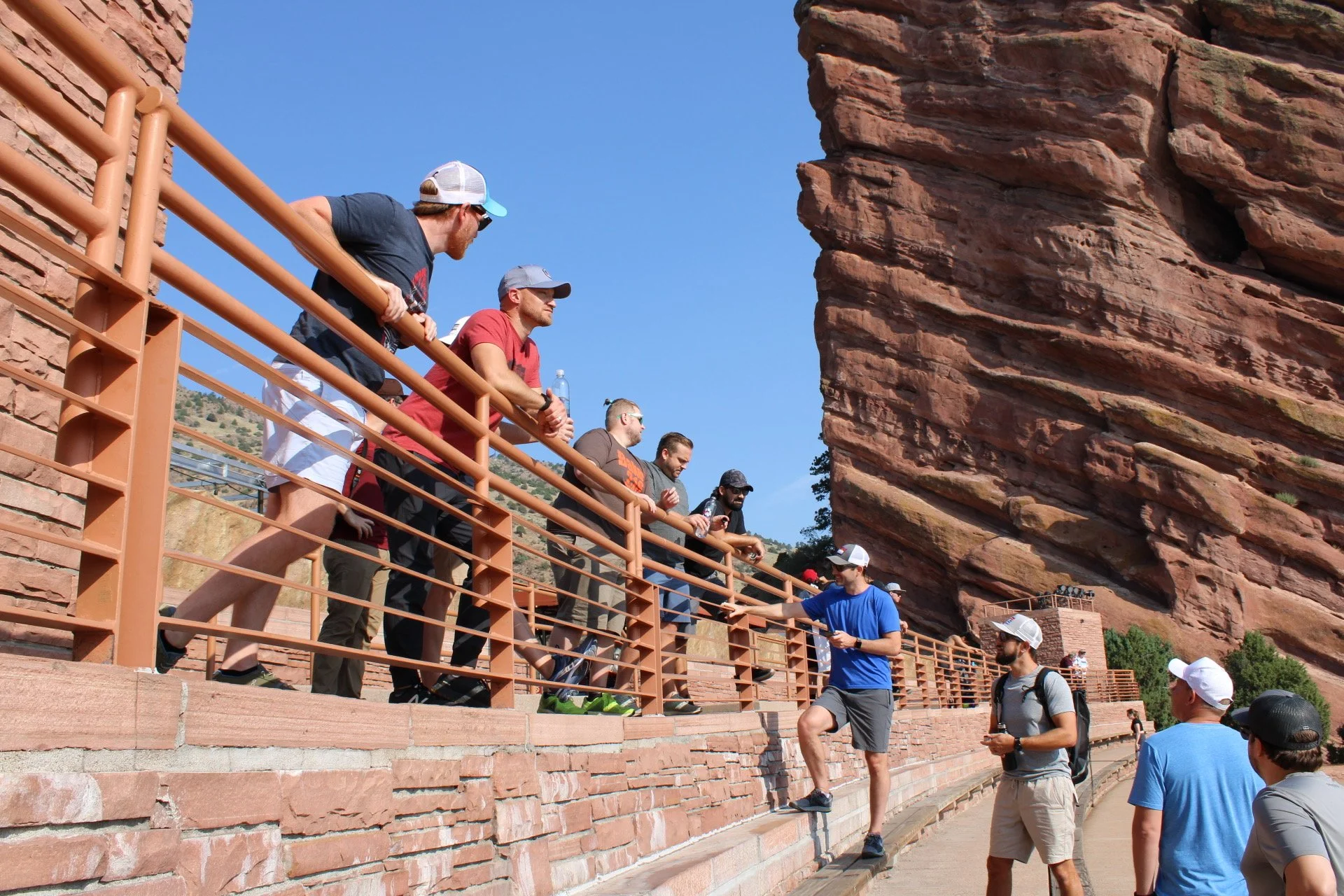 Group of people at the Garden of the Gods park, with large red rock formations in the background, some standing on a brick pathway and others near a railing, engaging in conversation on a sunny day.