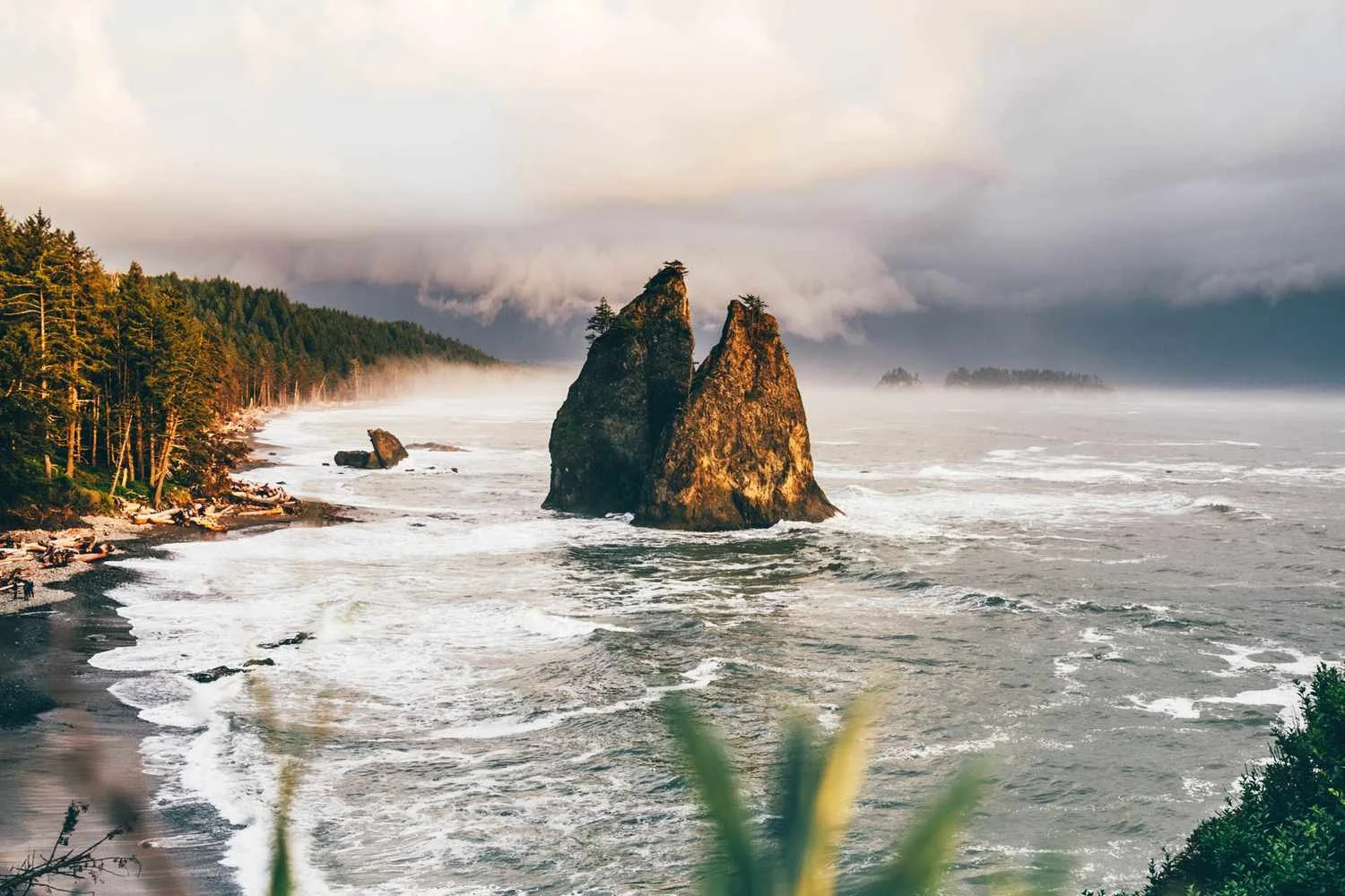 Scenic view of Oregon coast with large sea stacks, forested shoreline, and waves crashing against rocks, under cloudy sky.