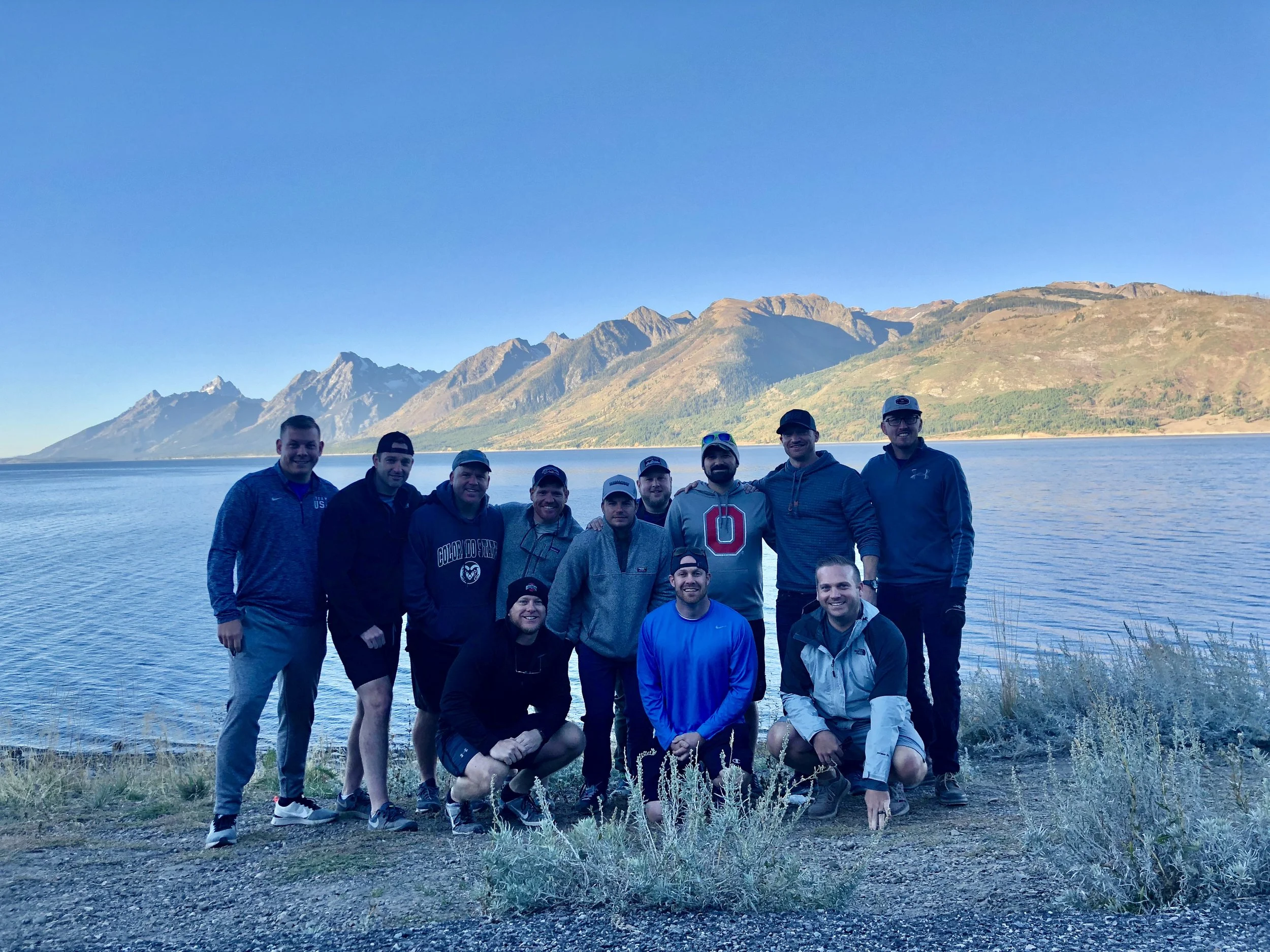 A group of eleven men posing by a large lake with a mountain range in the background, under a clear blue sky.