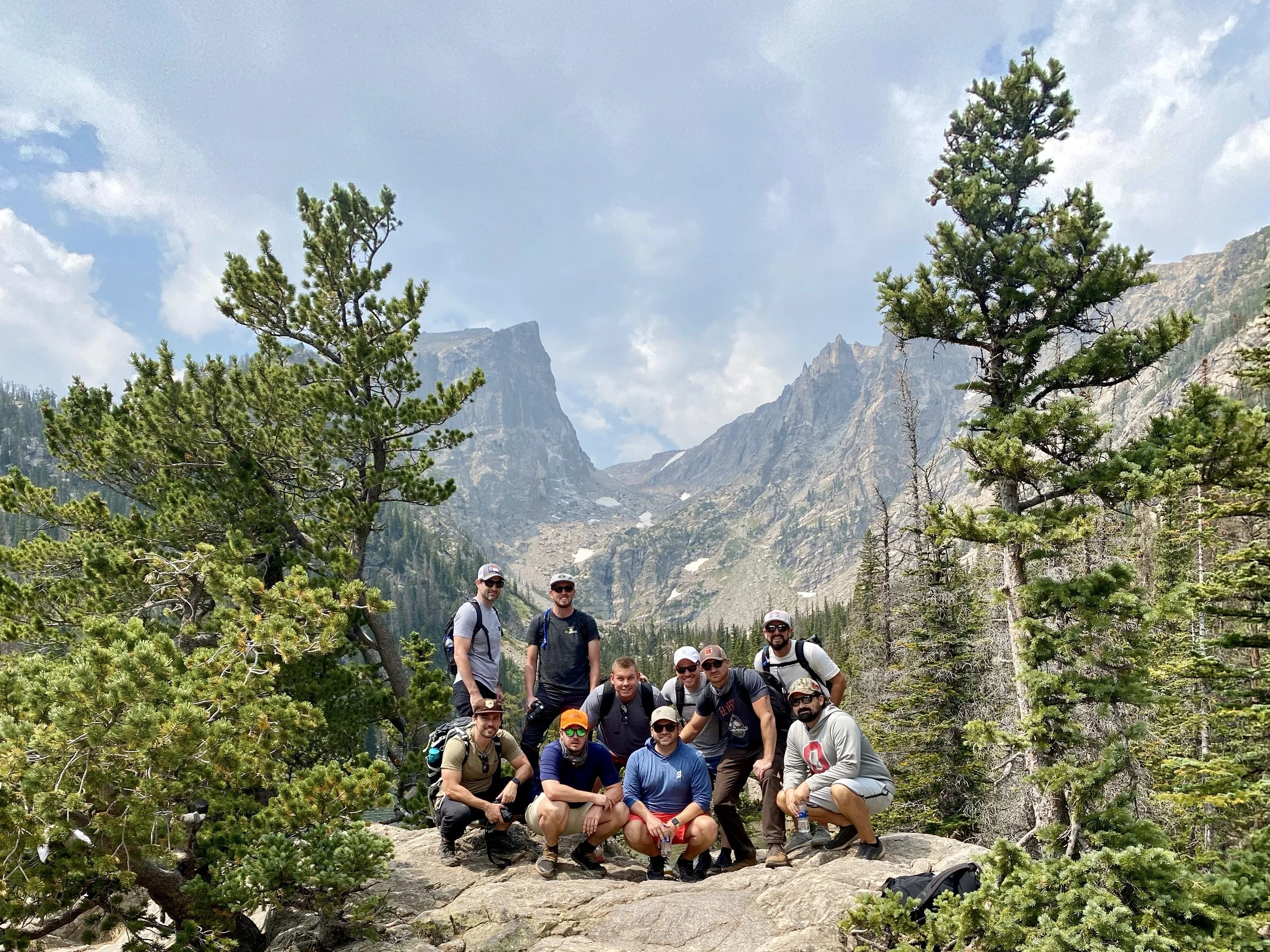 Group of nine hikers posing on a large rock in a mountainous, forested area with tall pine trees and rugged mountain peaks in the background.