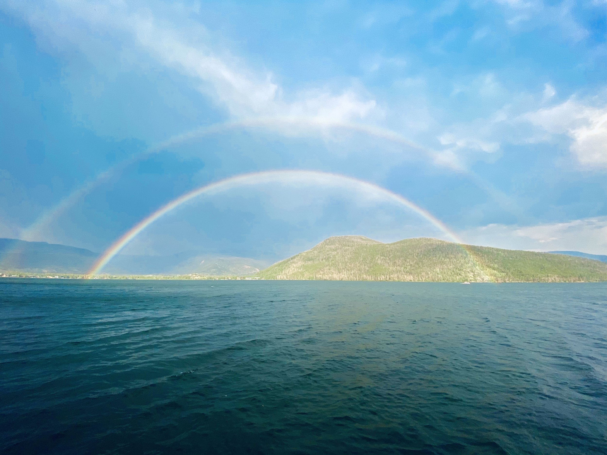 A double rainbow over a body of water with a mountainous landscape in the background under a partly cloudy sky.