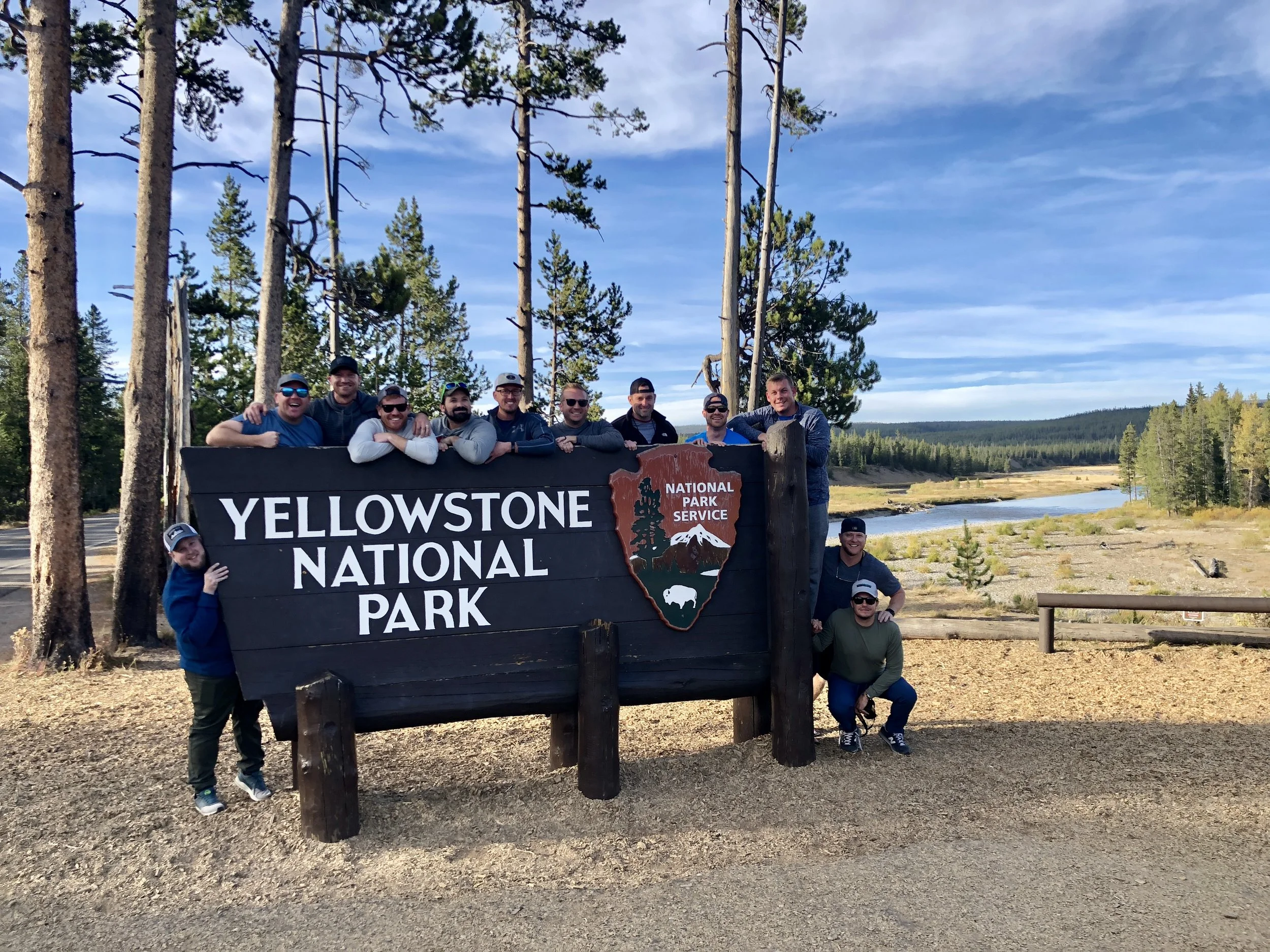 A group of eleven people posing behind a large sign that reads 'Yellowstone National Park' at Yellowstone National Park, with trees, a river, and a clear sky in the background.