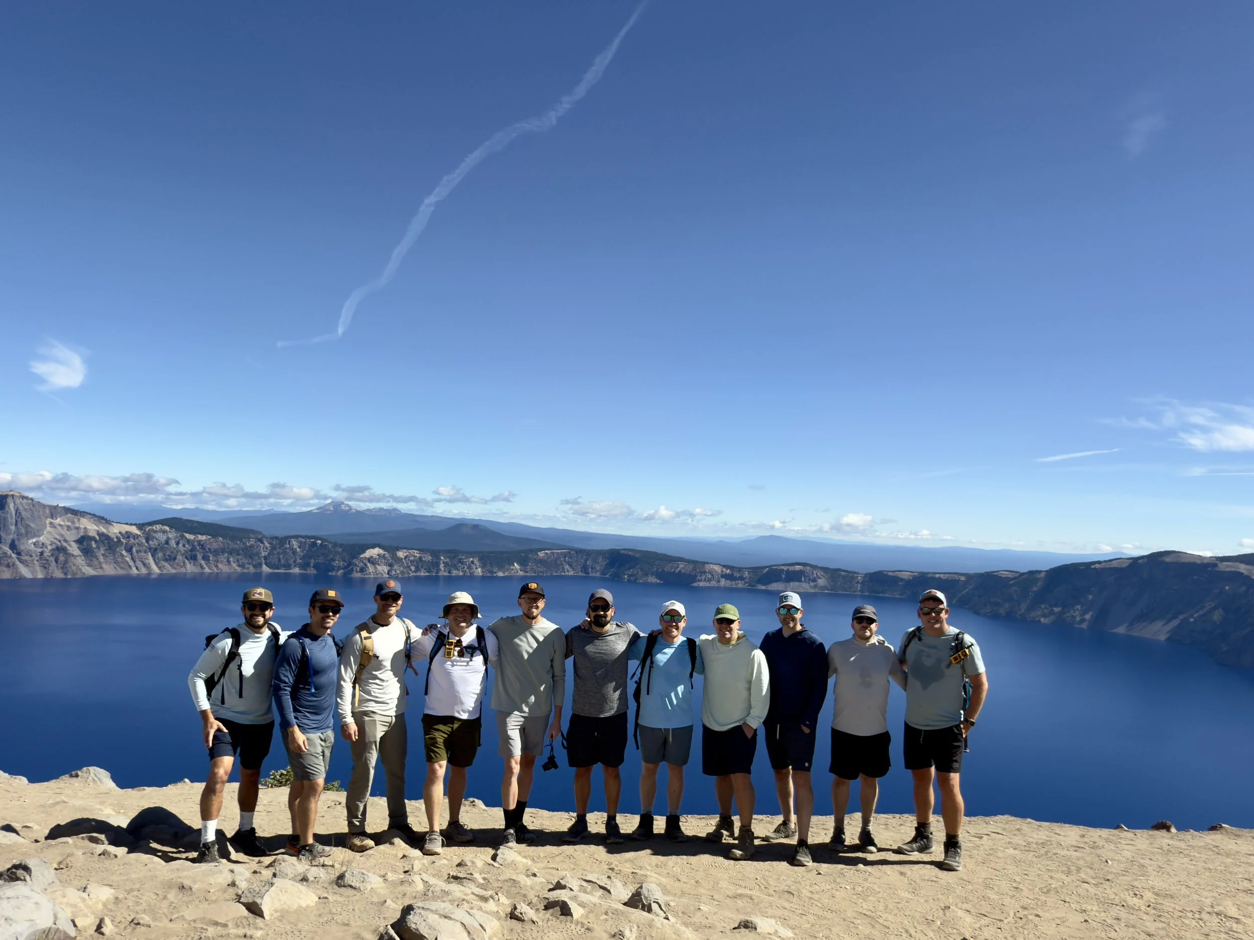 A group of eleven people standing on rocky terrain near a large blue lake with mountains in the background under a clear blue sky.