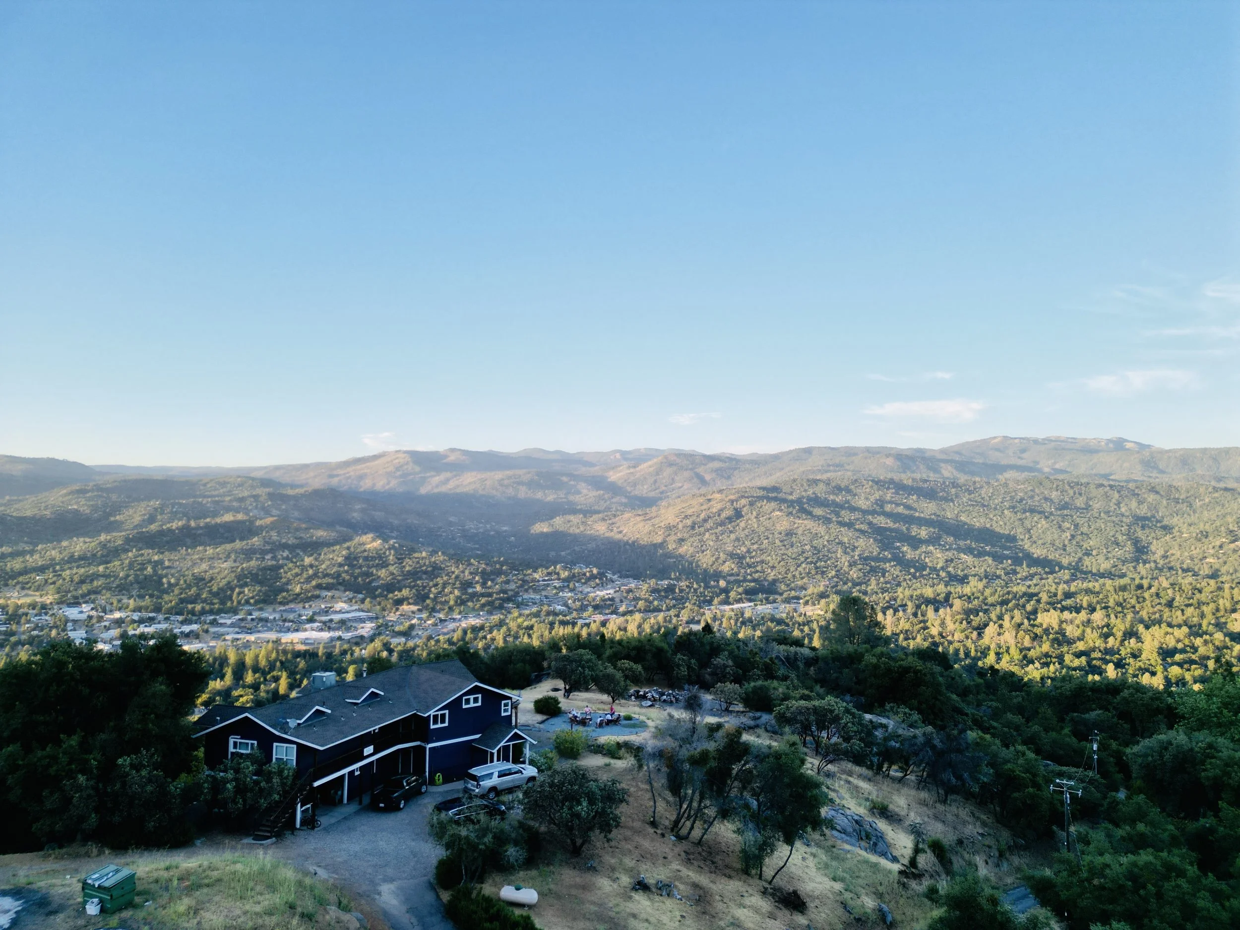 A landscape view of rolling hills and mountains under a clear blue sky, with a house and trees in the foreground.