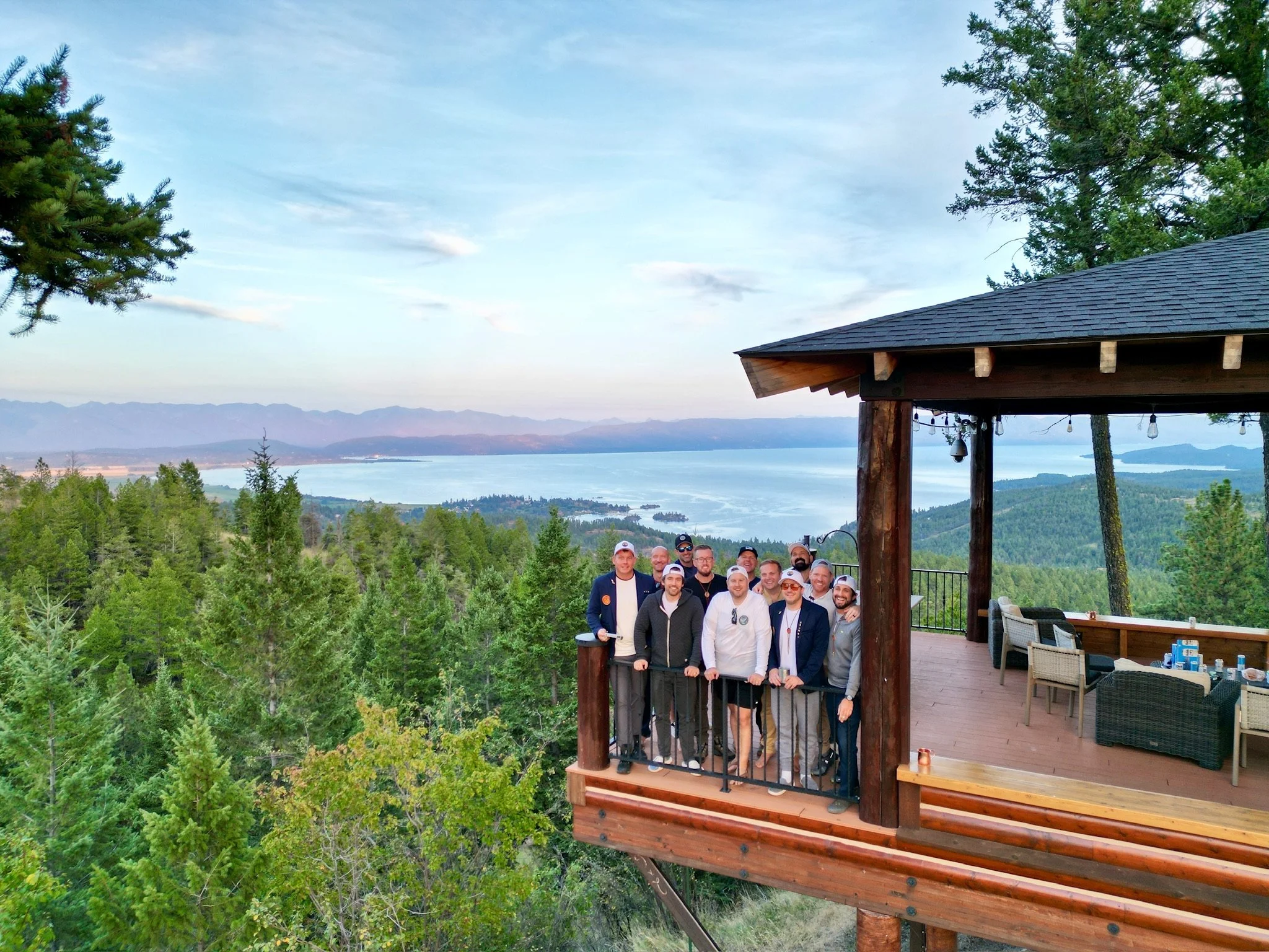 Group of people standing on a wooden balcony overlooking a forested landscape and lake with mountains in the background.