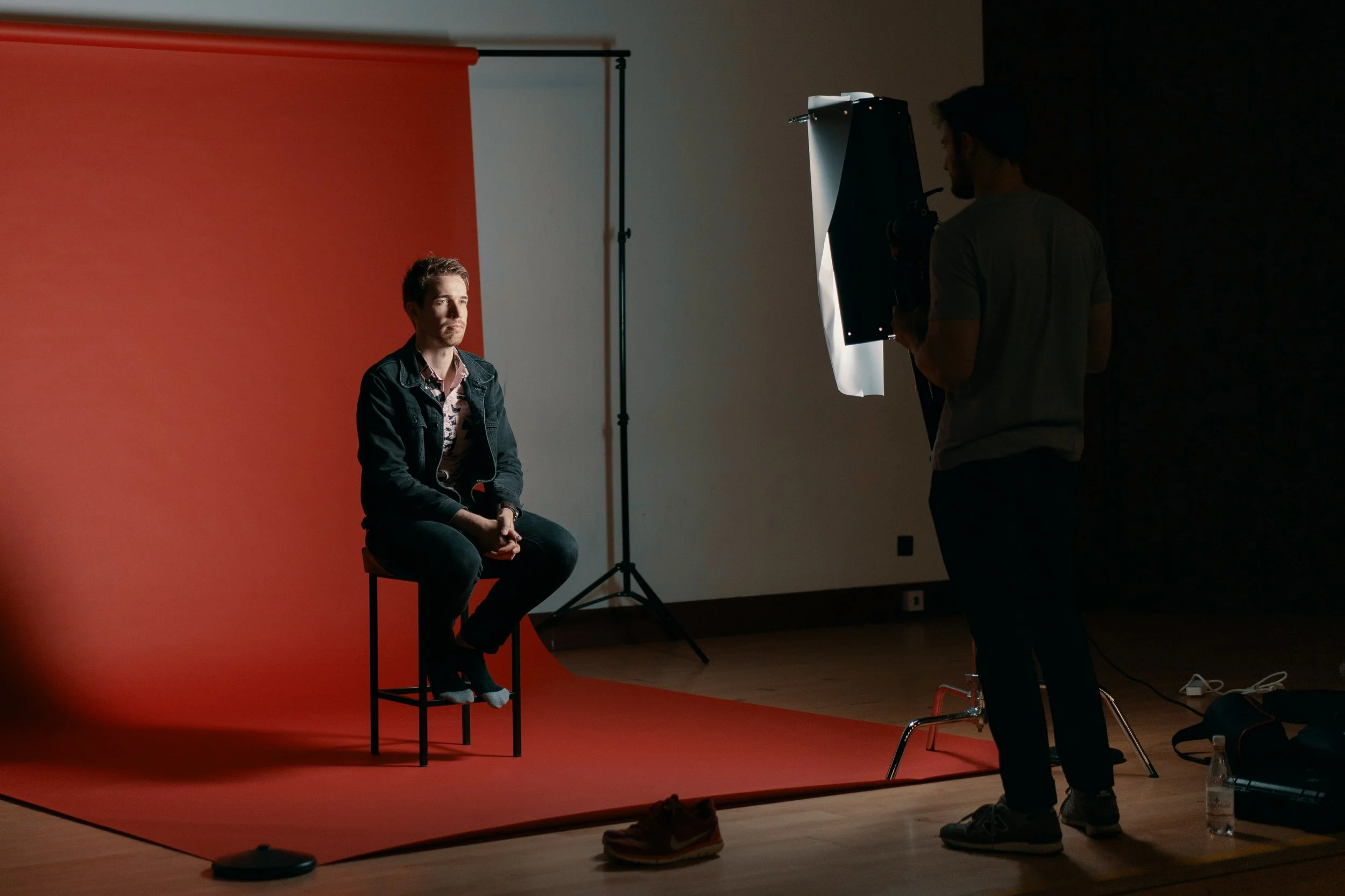 A man sitting on a stool in front of an orange backdrop during a photography session, with a photographer adjusting a light in a darkened studio.