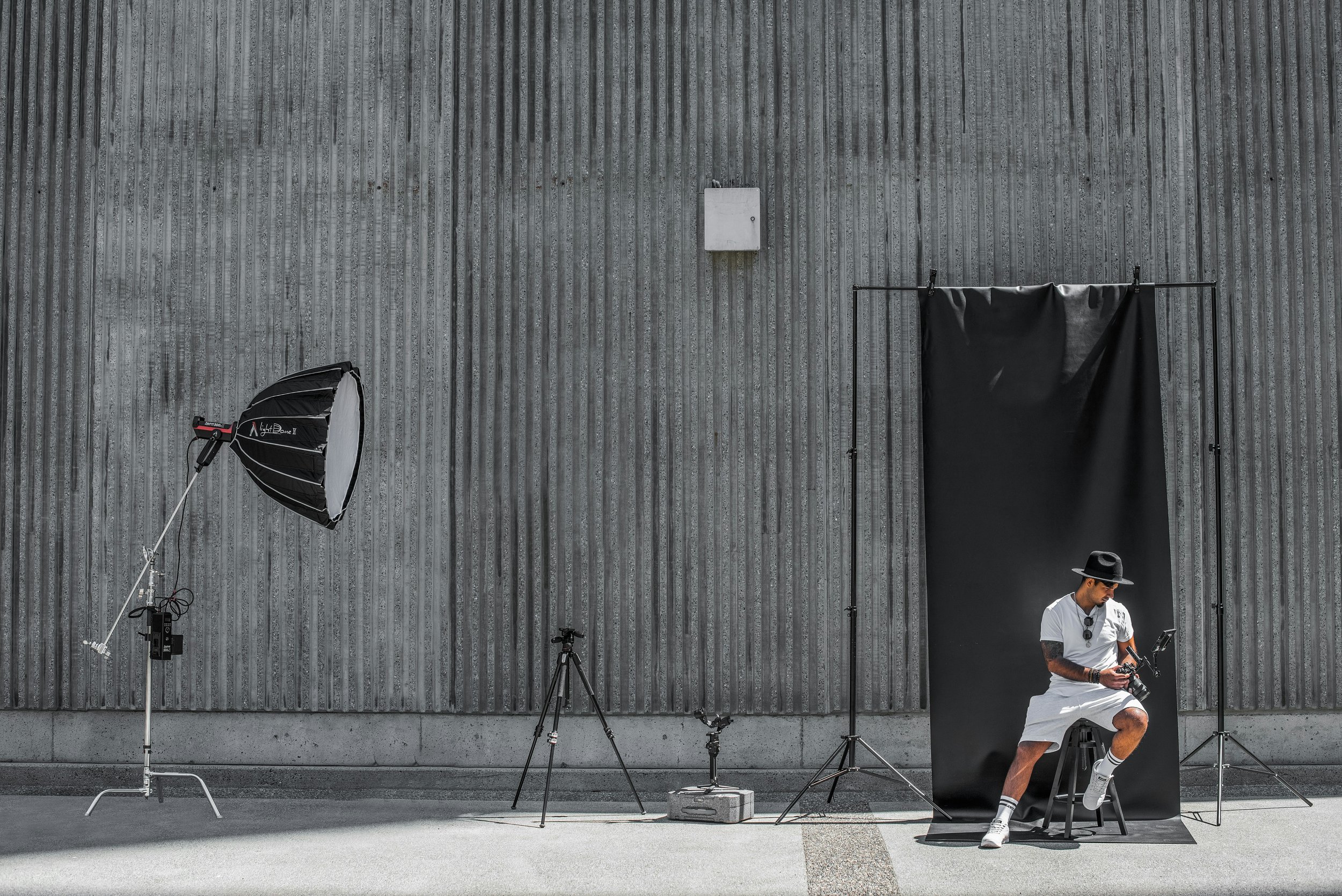 Man in white outfit with black hat and tattoos sitting on a stool in a photo studio with black backdrop and professional lighting equipment against a gray textured wall.
