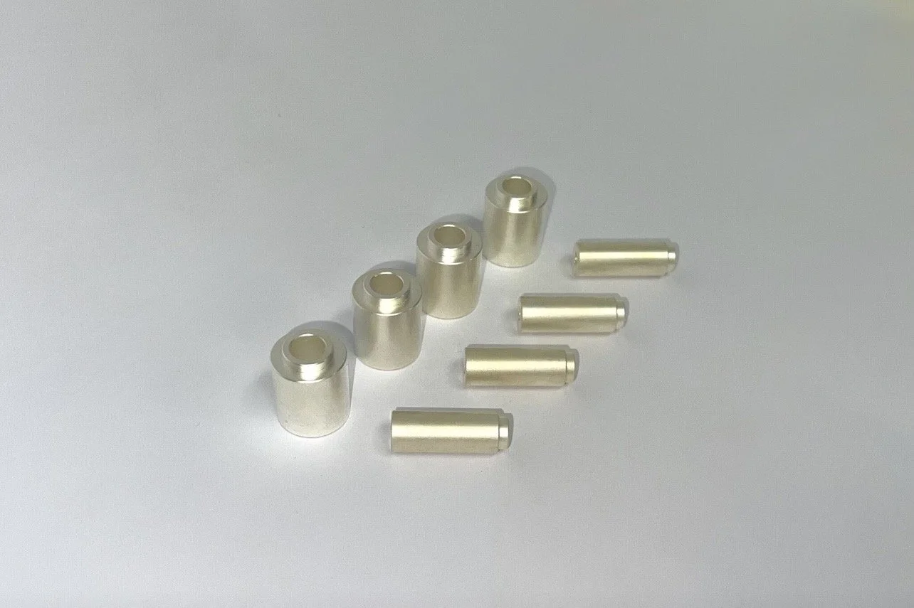 Six small cylindrical metal components with a hollow center and six smaller solid metal pins, all arranged on a plain light-colored surface.