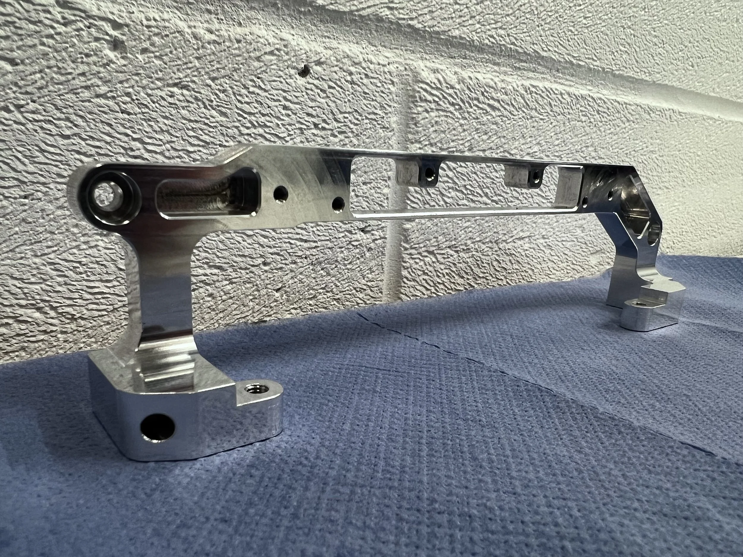 A shiny metallic car part with multiple holes, mounted on a textured blue surface against a rough white wall.