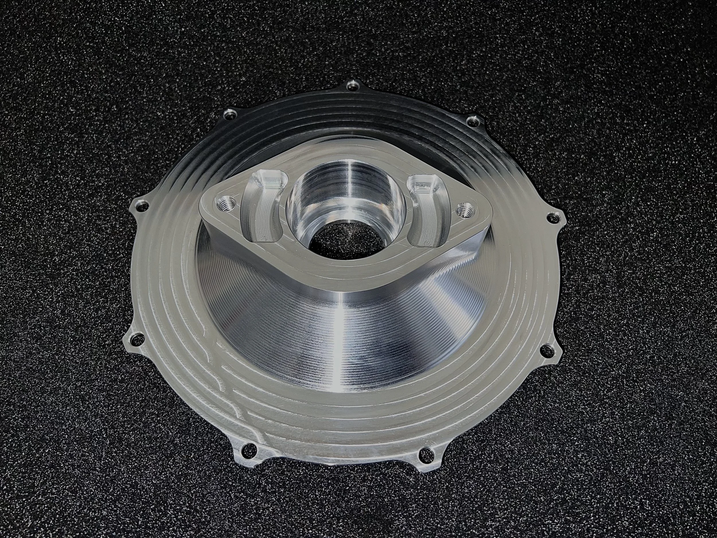 A metallic automotive or mechanical component with a central circular hole, surrounded by a conical surface and mounting holes, resting on a textured black surface.