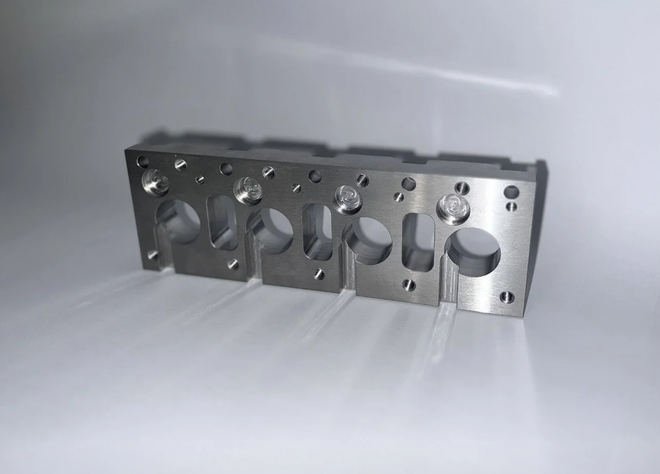 A metallic engine cylinder head with multiple holes and chambers, placed on a white surface.