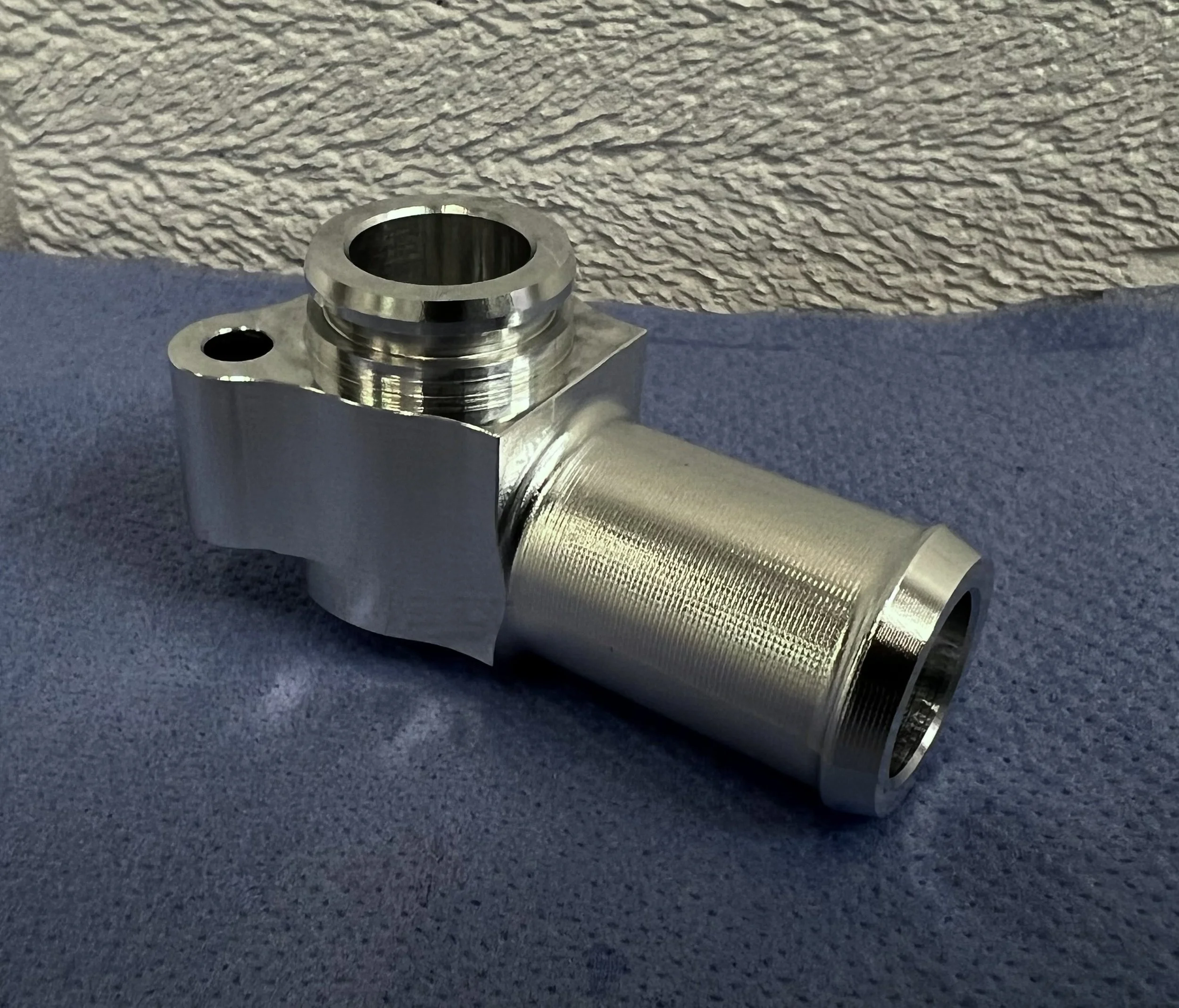 A close-up of a metallic mechanical part with a textured surface and a hollow cylindrical section, placed on a blue fabric surface with a textured wall in the background.