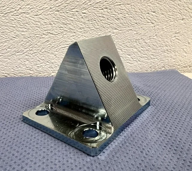 A metallic, pyramid-shaped mechanical component with a threaded hole in the center, mounted on a perforated metal base.