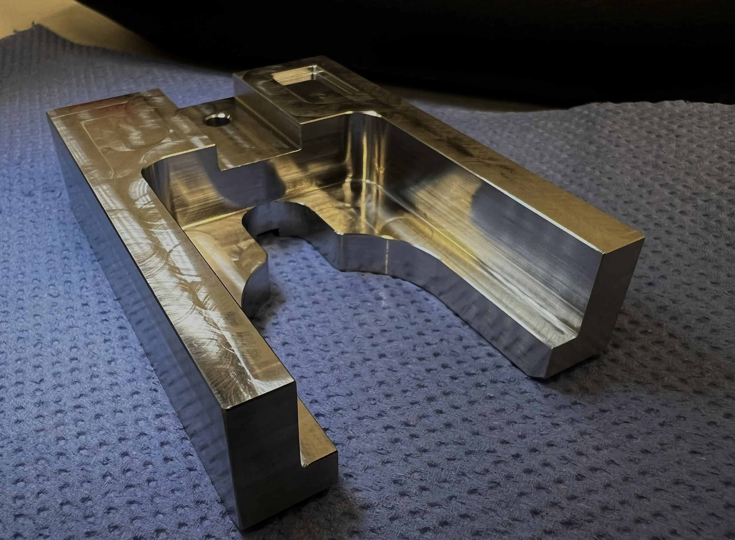 A metallic, machined part with a rectangular shape, rounded edges, and a curved cut-out section, resting on a textured gray surface.