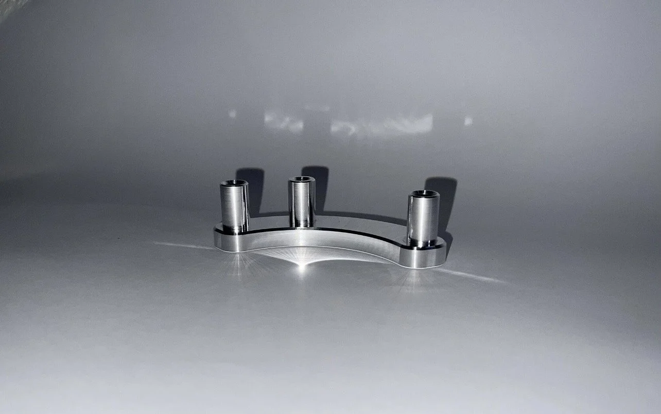 A metallic bracket with four cylinders attached, placed on a smooth flat surface, casting shadows on a gray background.