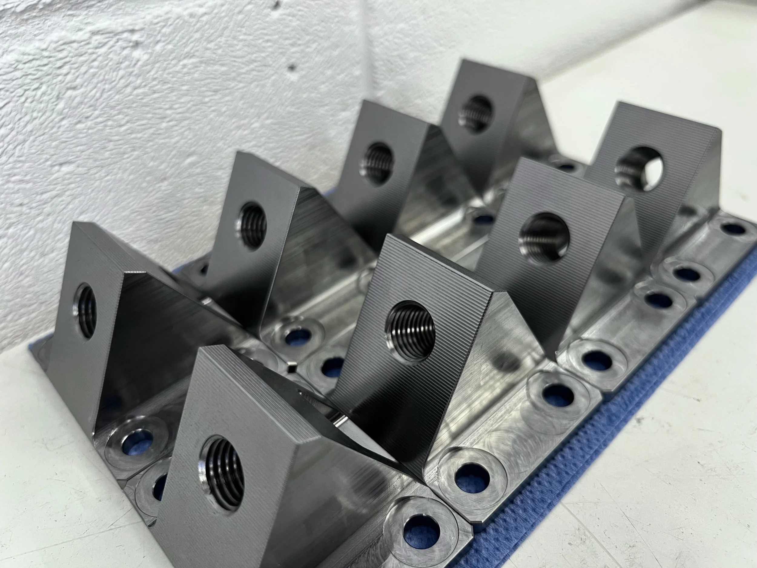 Set of metallic brackets with threaded holes arranged on a white surface against a textured wall.