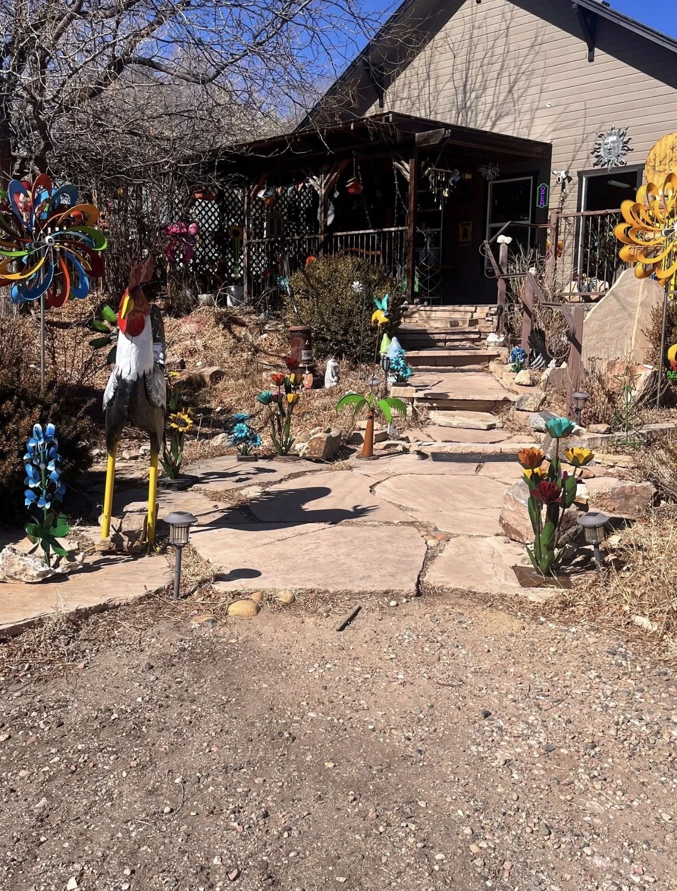 Front yard decorated with colorful wind spinners, metal flowers, and garden ornaments, leading to a house with stairs and a porch.