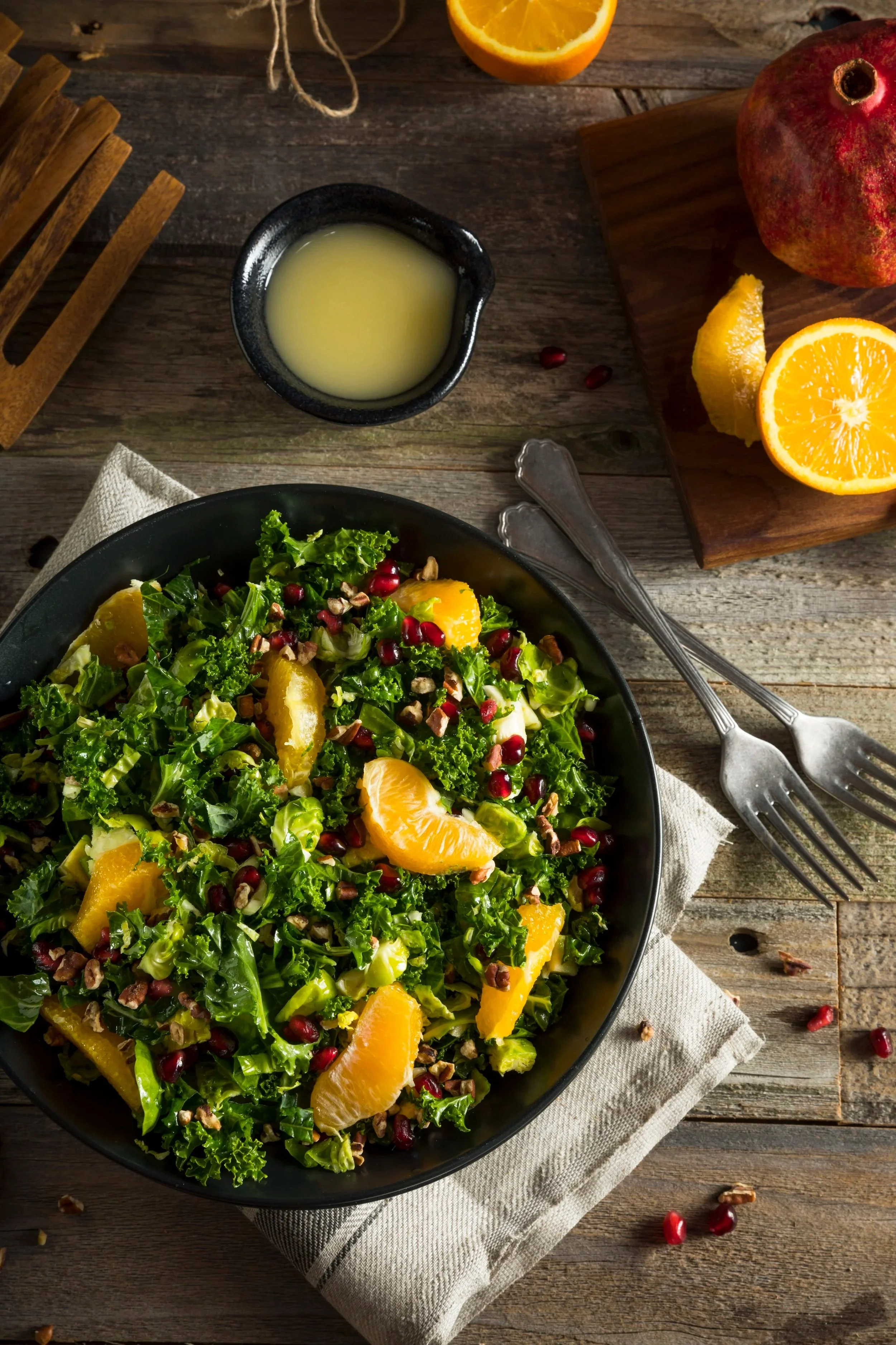 A fresh green salad with orange slices, pomegranate seeds, and chopped nuts in a black bowl on a rustic wooden table, with a pomegranate, orange halves, and a lemon wedge nearby.