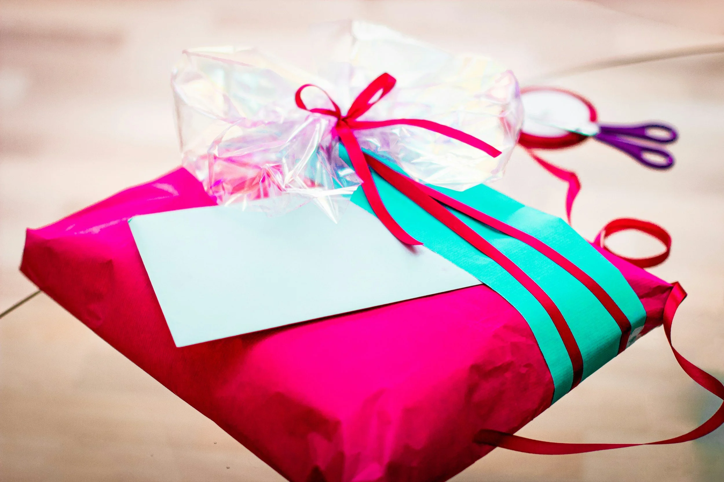 A brightly wrapped pink gift box with a light blue ribbon and a white blank tag, topped with a clear plastic bag tied with a pink ribbon, on a wooden surface.