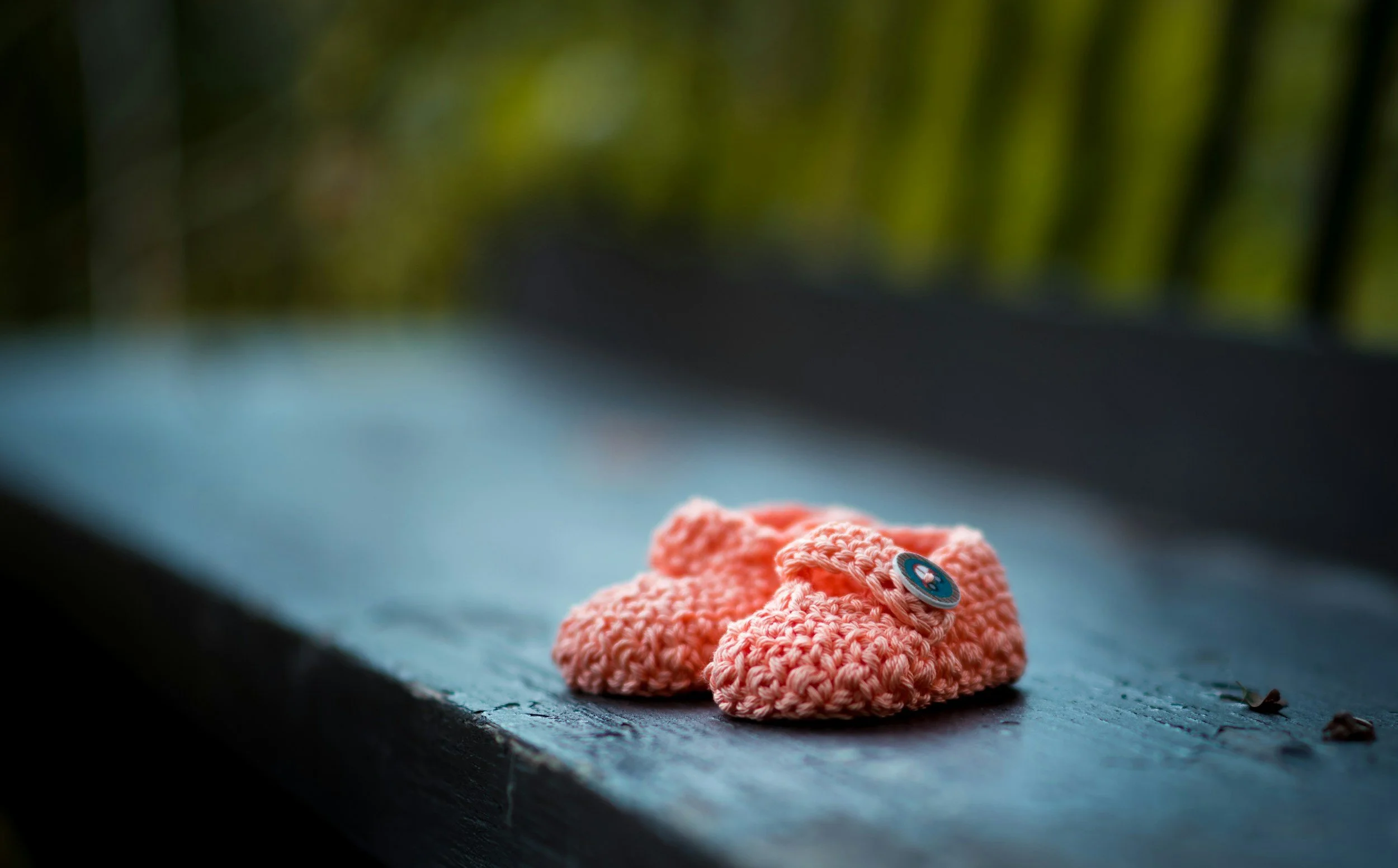 A pair of small, pink, crocheted baby shoes with a button detail on a weathered dark blue wooden surface outdoors.