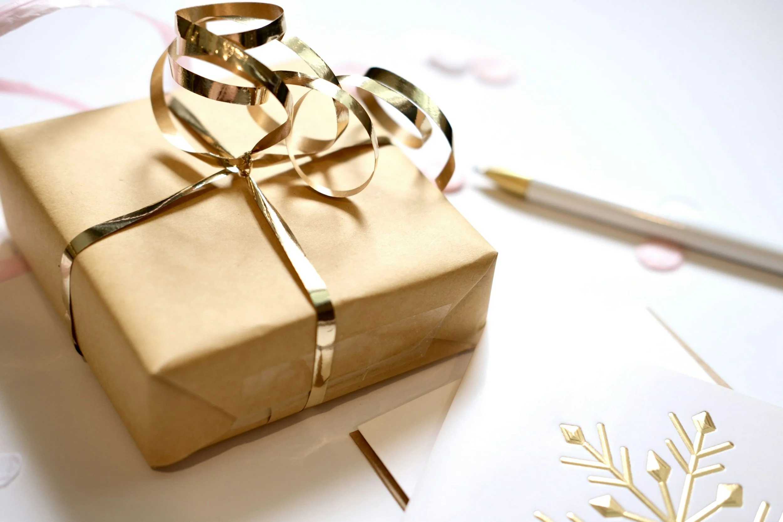 A gold-wrapped gift box with a gold ribbon and bow, surrounded by pink confetti, next to a white pen and a greeting card with a gold snowflake design.