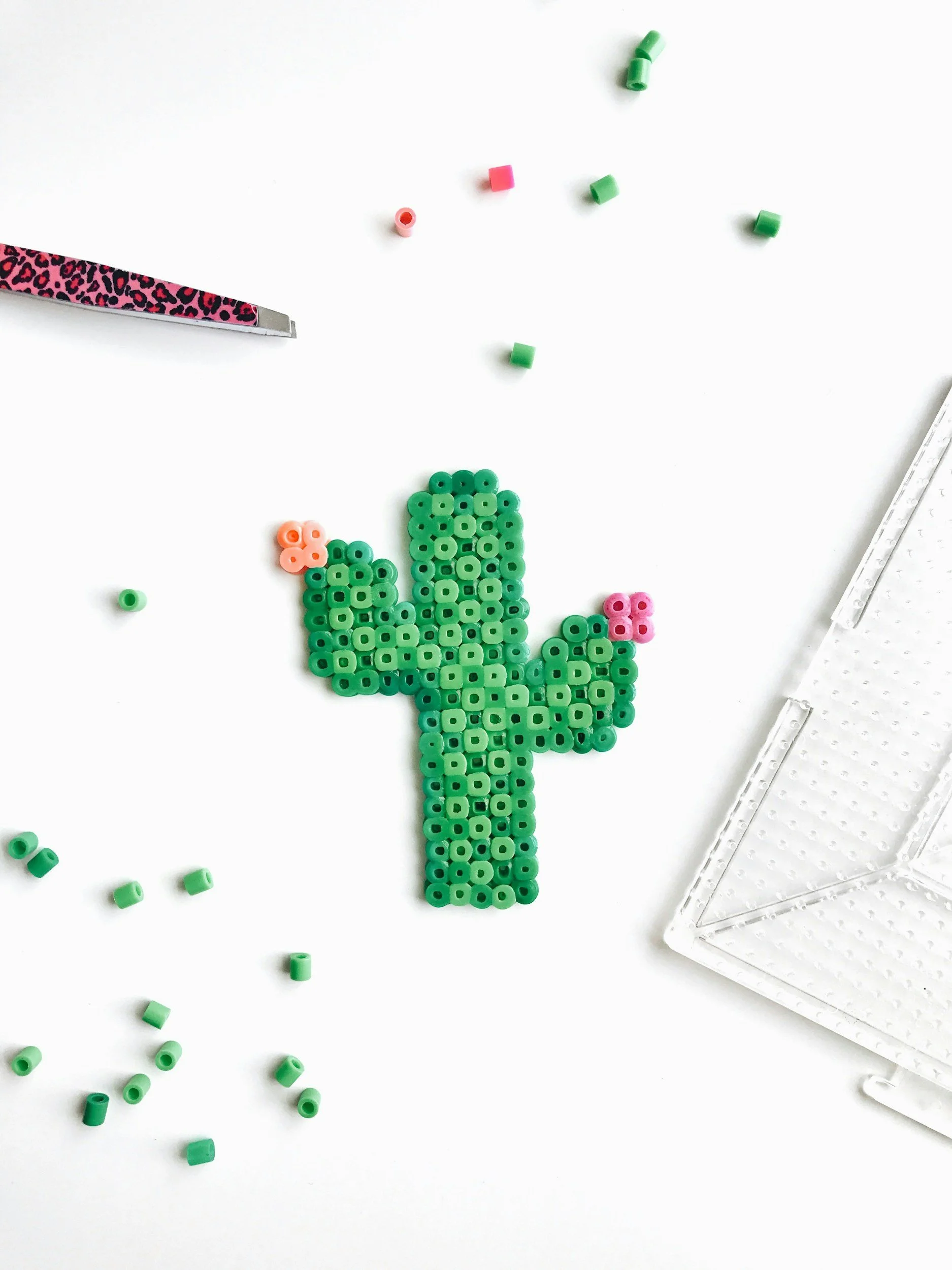 Green cactus beaded art with pink and peach flowers, scattered beads, a patterned craft knife, and a clear storage container on a white surface.