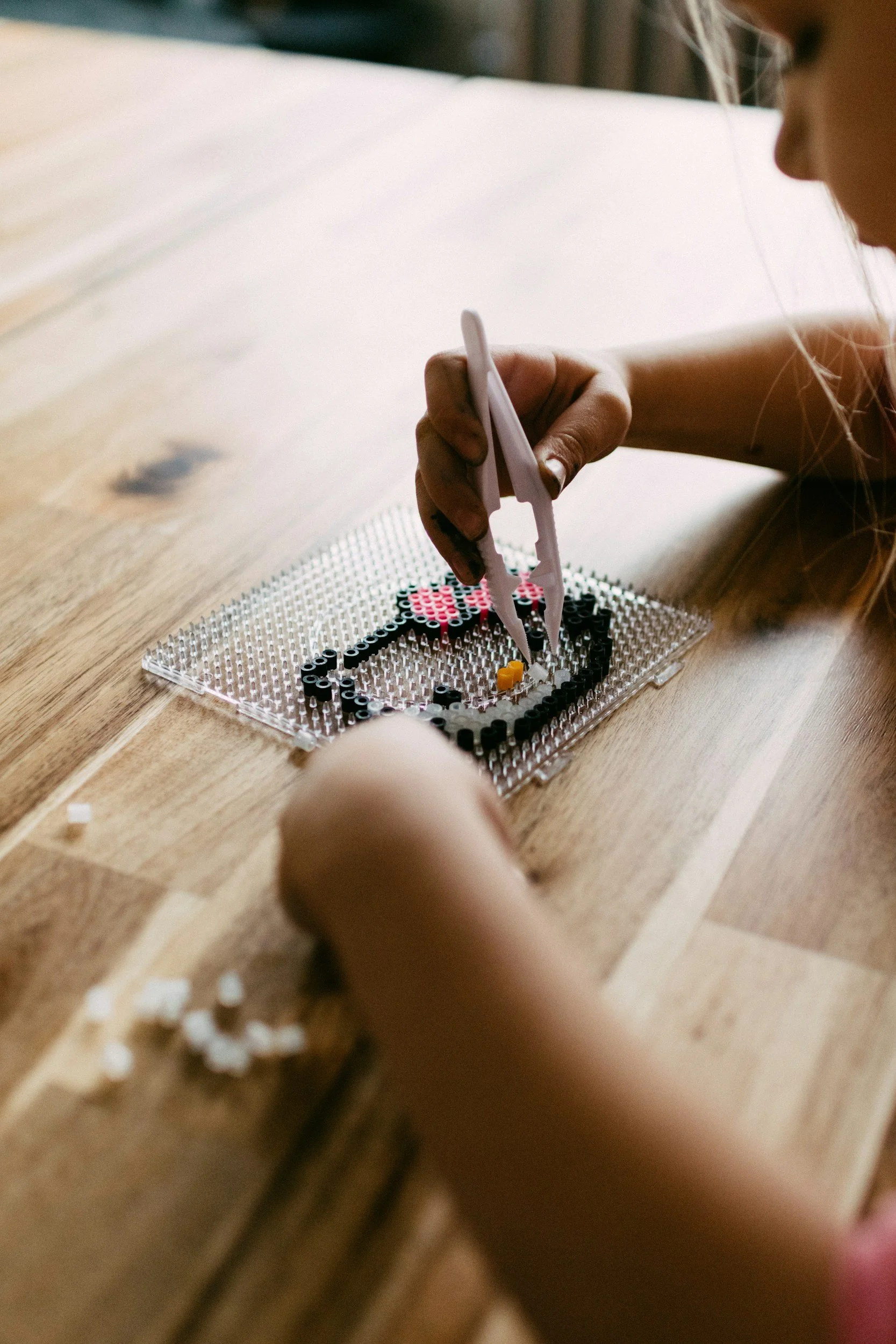 Child uses tweezers to place small beads onto a pegboard to create a mosaic design, sitting at a wooden table.