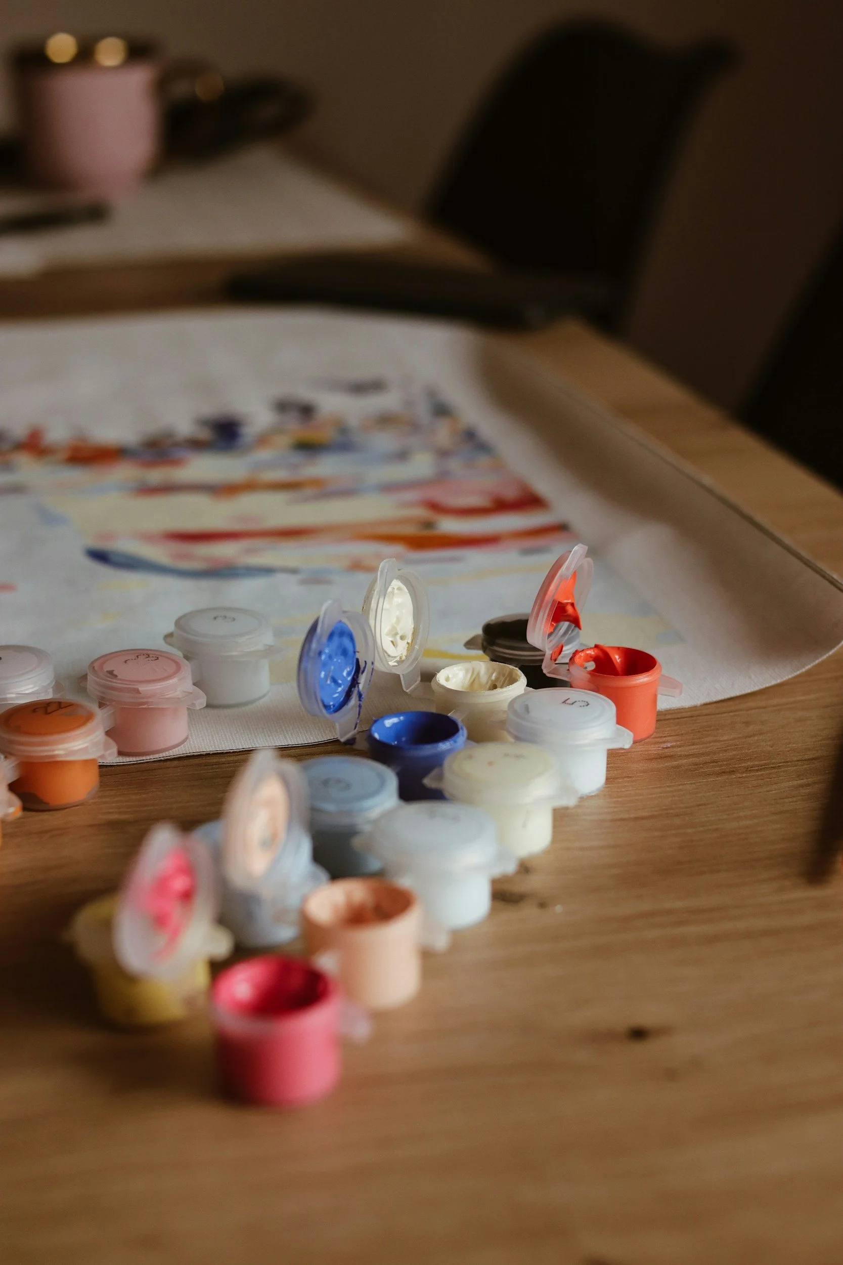 Open paint color containers arranged on a wooden table next to a canvas with paint strokes.