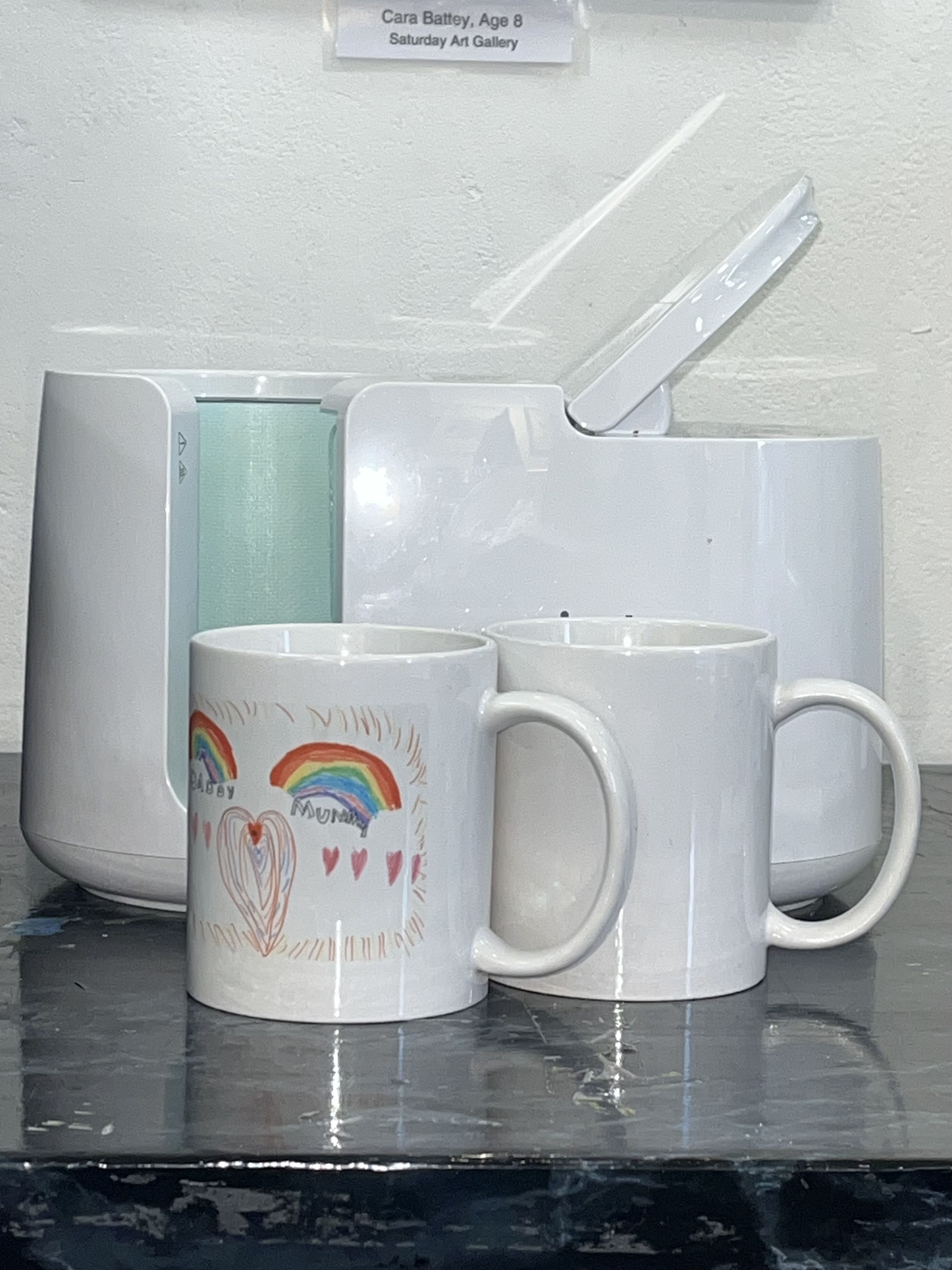 Two white coffee mugs with children's rainbow drawings and hearts on one mug, placed in front of a white ice shaver machine on a black countertop.