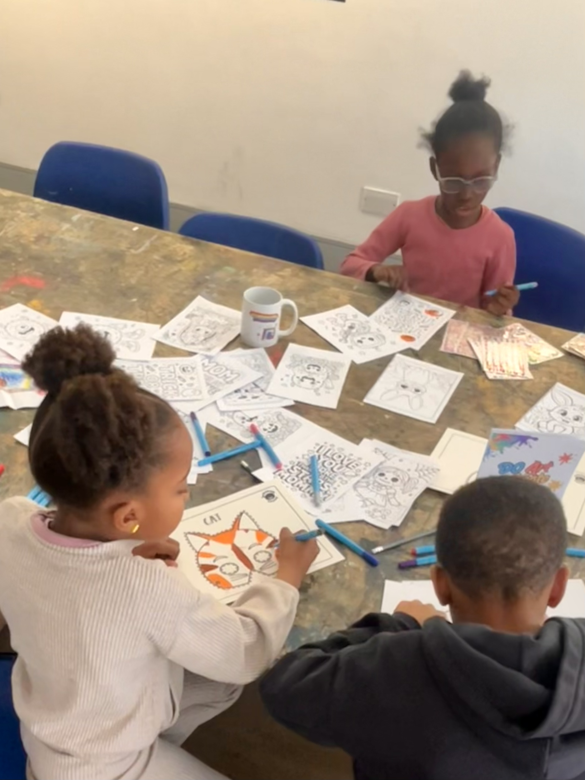 Three children are coloring and drawing at a table filled with coloring pages, markers, and a mug, in a classroom setting.