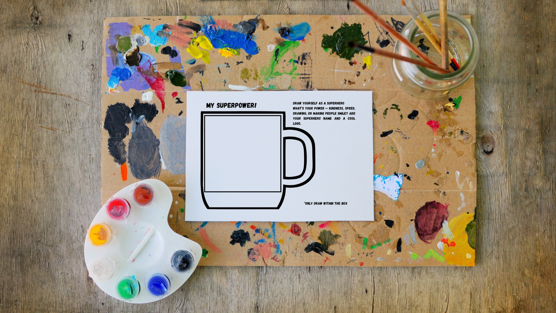 A creative workspace with a paint palette containing various colors, a paint-covered workstation, and a drawing of a superhero mug on a printed activity sheet, surrounded by a jar of paintbrushes.