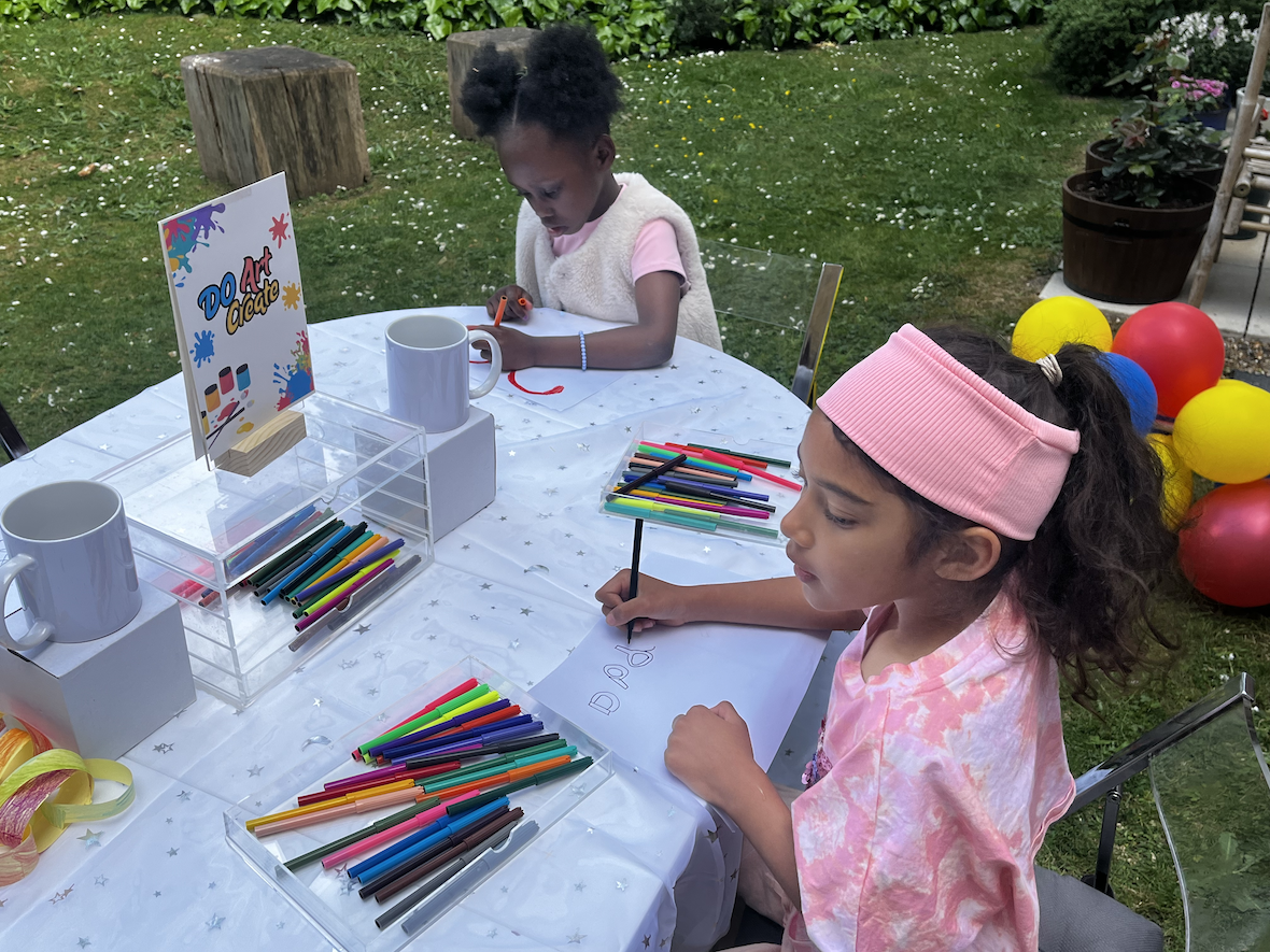 Two young girls sitting at a table outside with art supplies, coloring and drawing during a do art party, with balloons in the background.