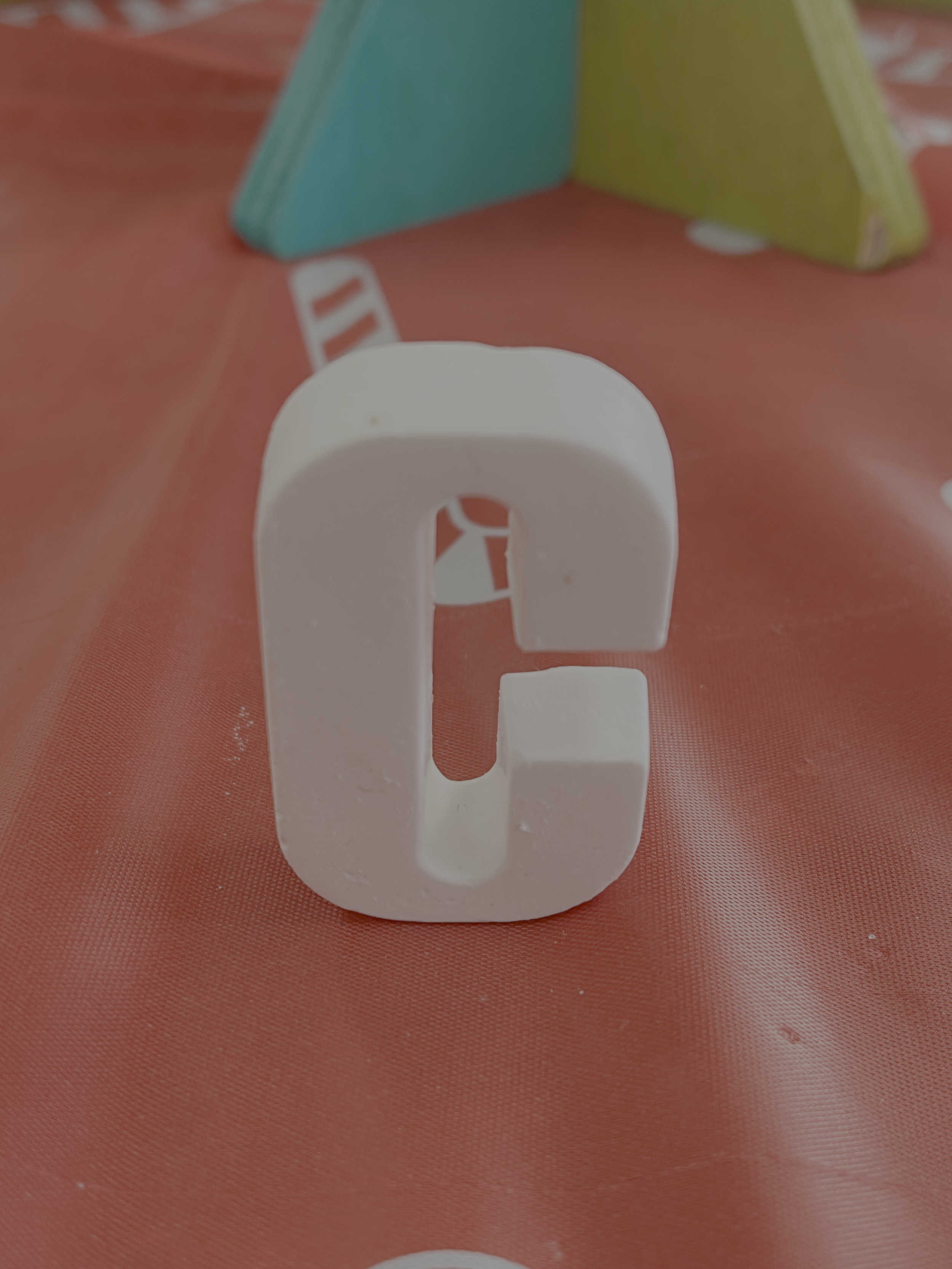 A white plaster 3D letter C on a pink surface with colourful small books in the background.